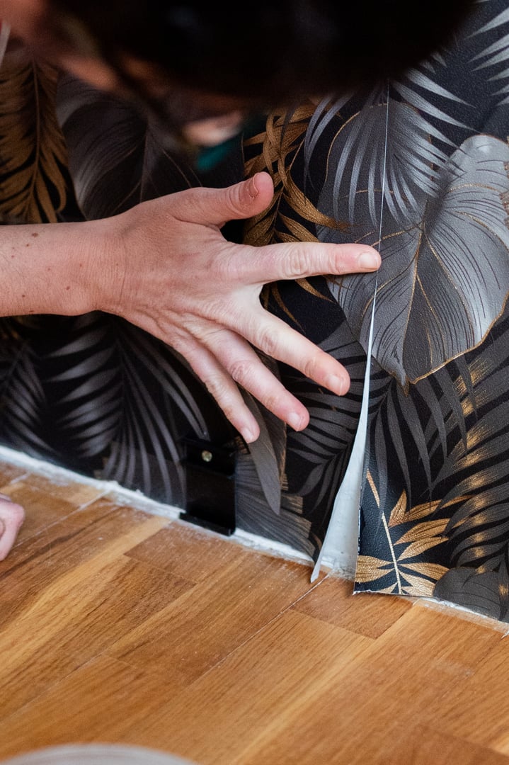Hand applying floral wallpaper along the edge of a wooden floor, showcasing a diy home improvement project