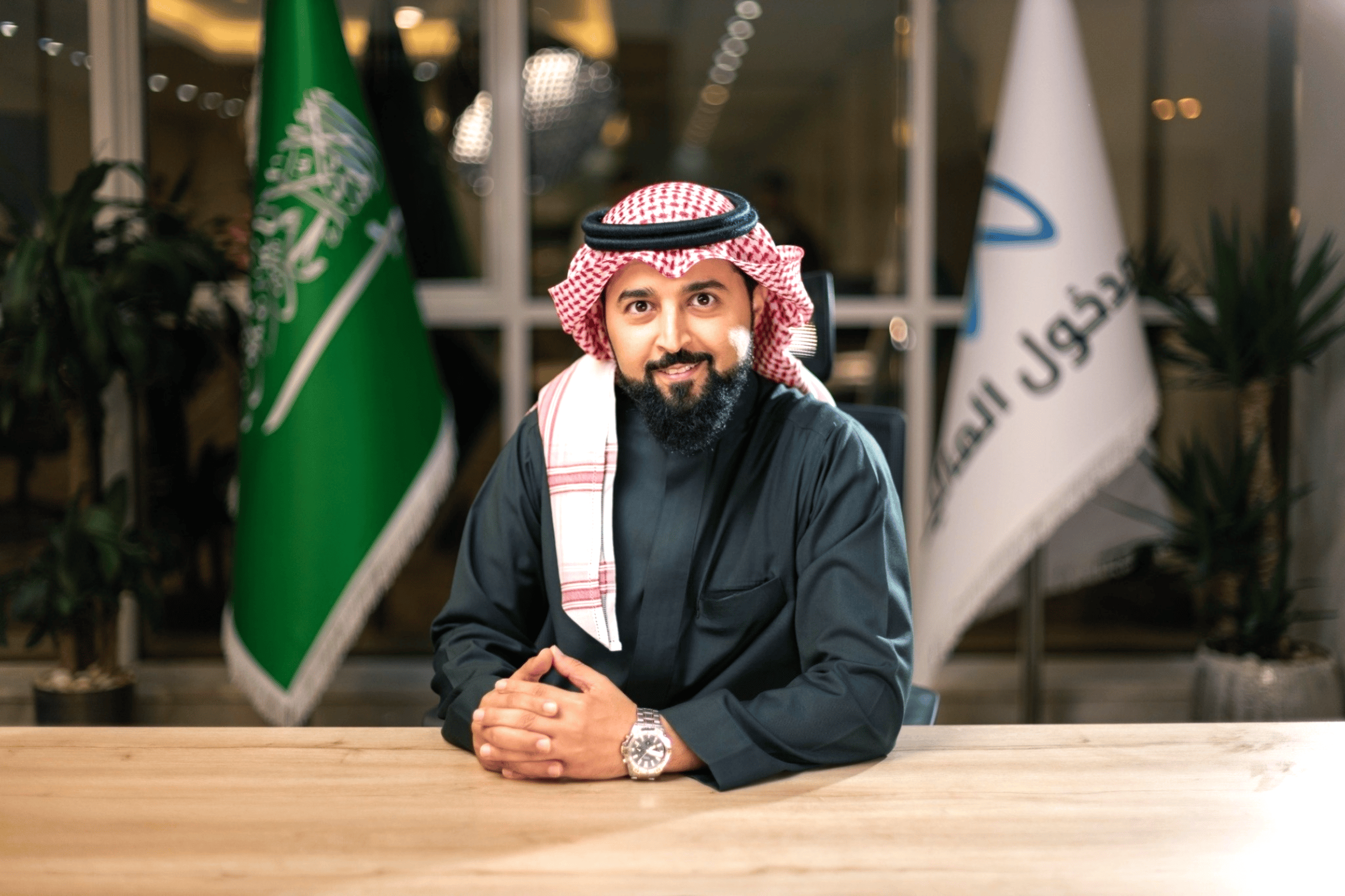 Man in traditional Saudi attire sits at a table with Saudi and logo flags in the background.