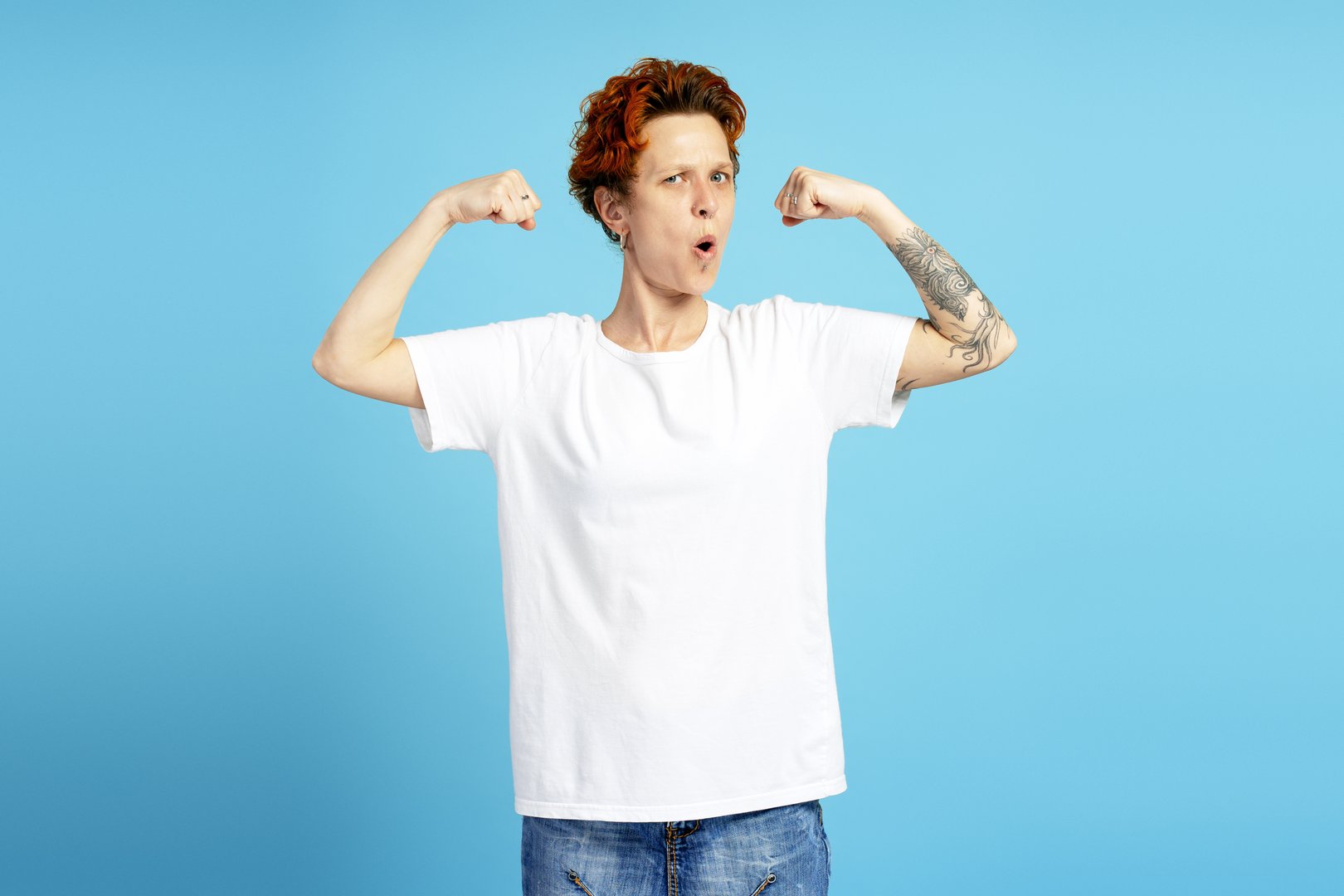 Surprised red-haired woman with arm tattoos demonstrating strength power, showing biceps muscles, isolated on blue. Individuality, diversity, and self-expression concept