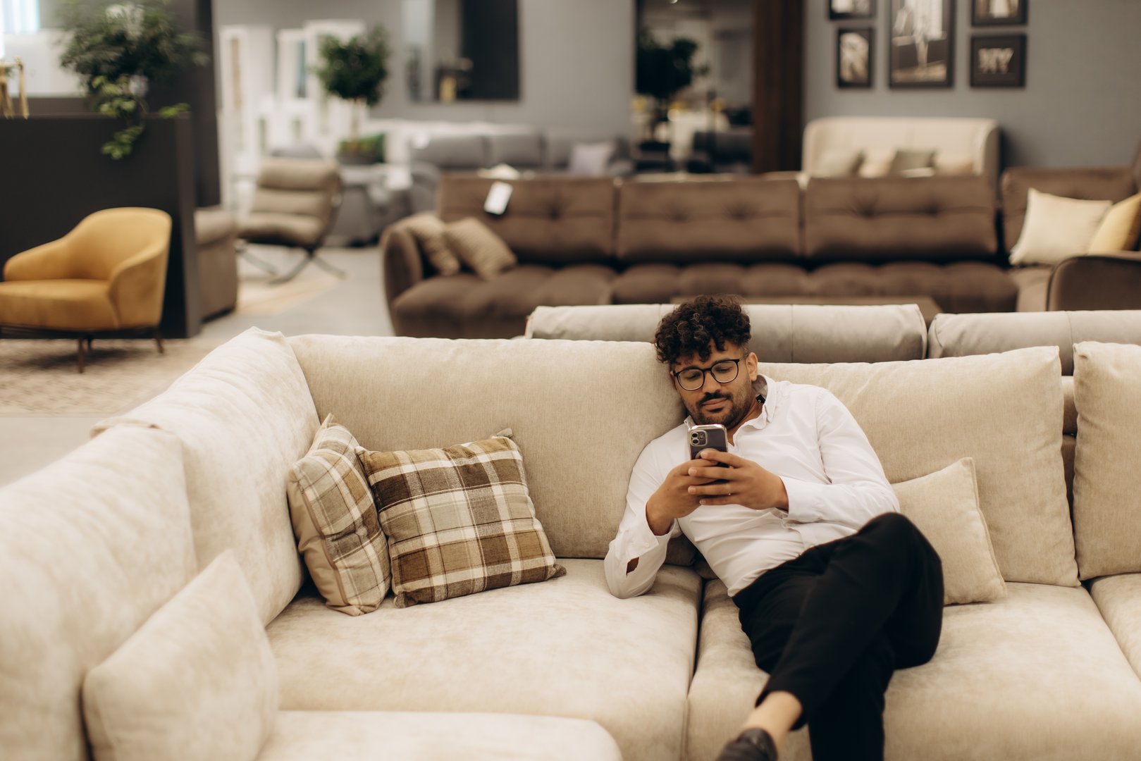 A man dressed in casual attire lounges on a cozy beige sofa, browsing his smartphone, within a stylish and modern furniture showroom featuring a variety of seating and decor options.