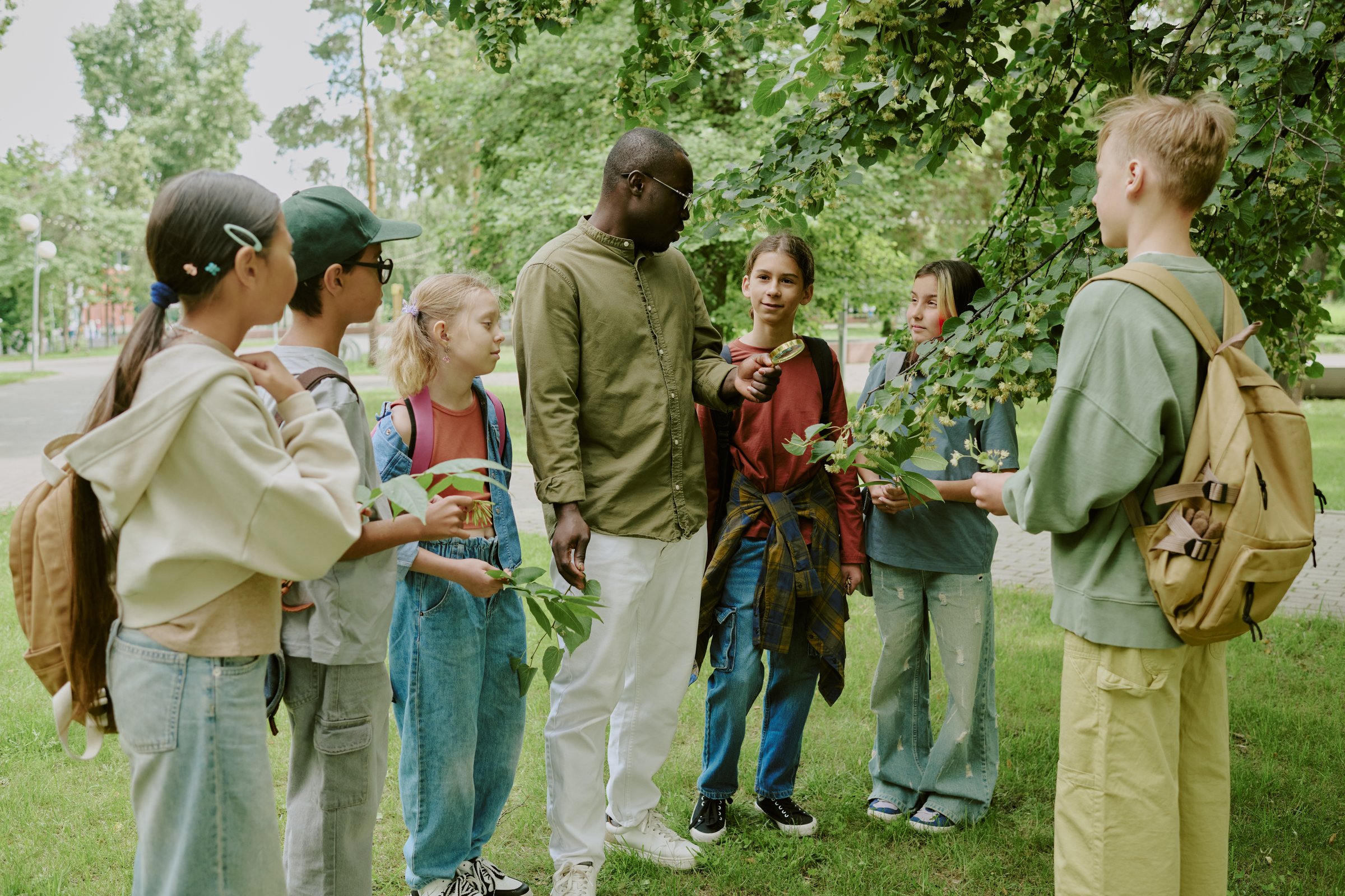 Group of diverse children and Black male teacher standing outdoors in park, teacher engaging students in nature lesson, children holding leaves and listening attentively during educational activity
