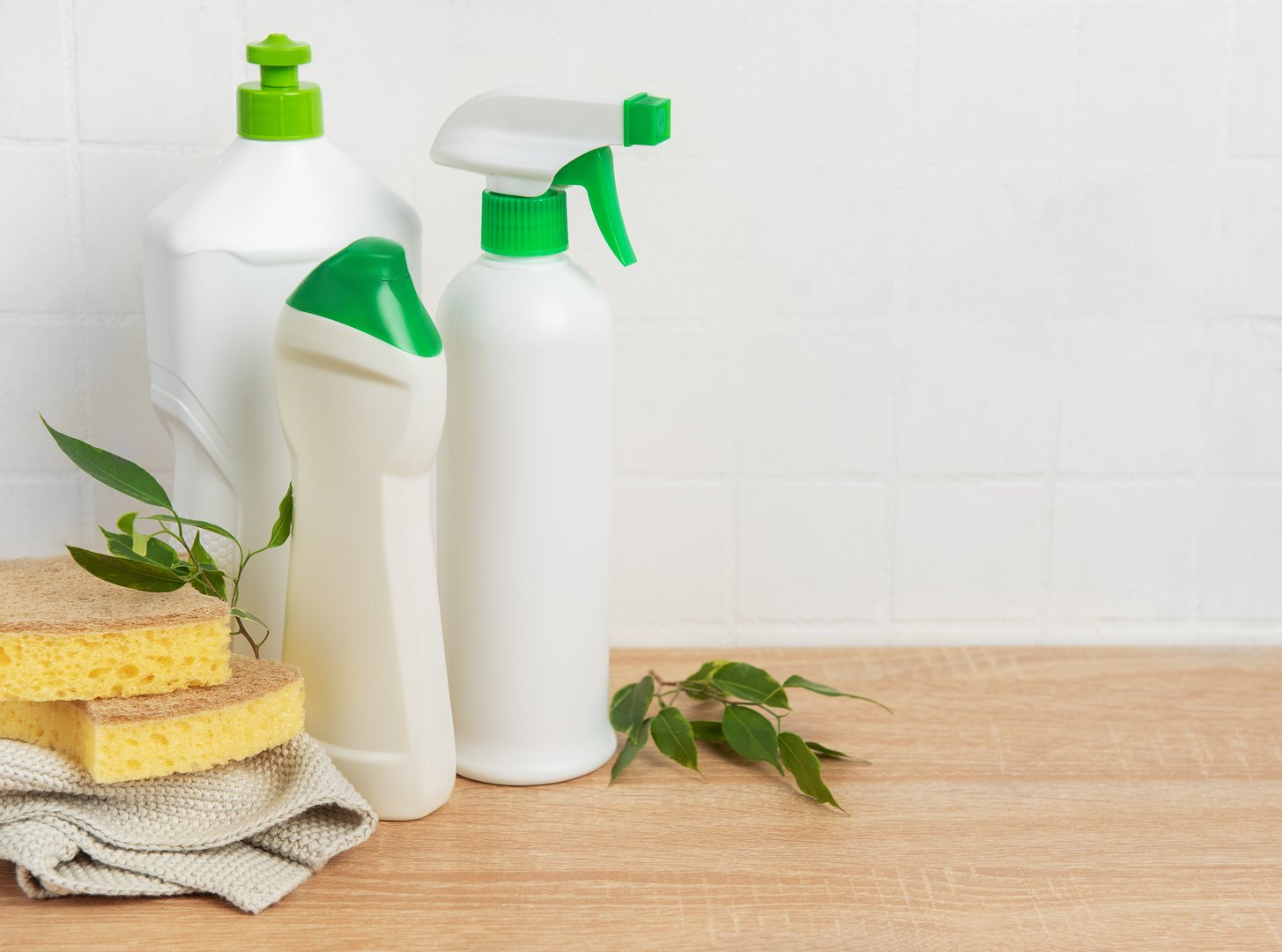 Eco friendly cleaning products, sponges, and green leaves are sitting on a wooden table against a white tiled wall, promoting sustainable cleaning practices