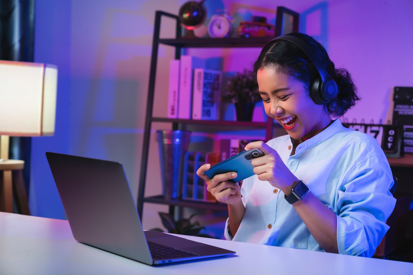 Excited Young Asian woman wearing headset and playing online game on smartphone at night modern house.