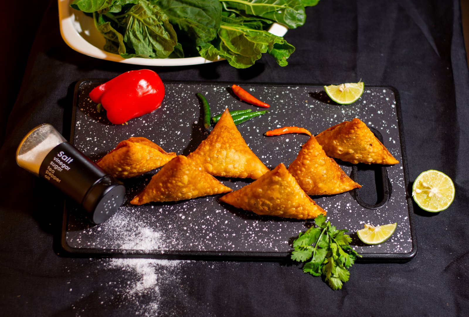 Samosas on a black cutting board with salt, red pepper, green chilies, cilantro, spinach leaves, and lime wedges.