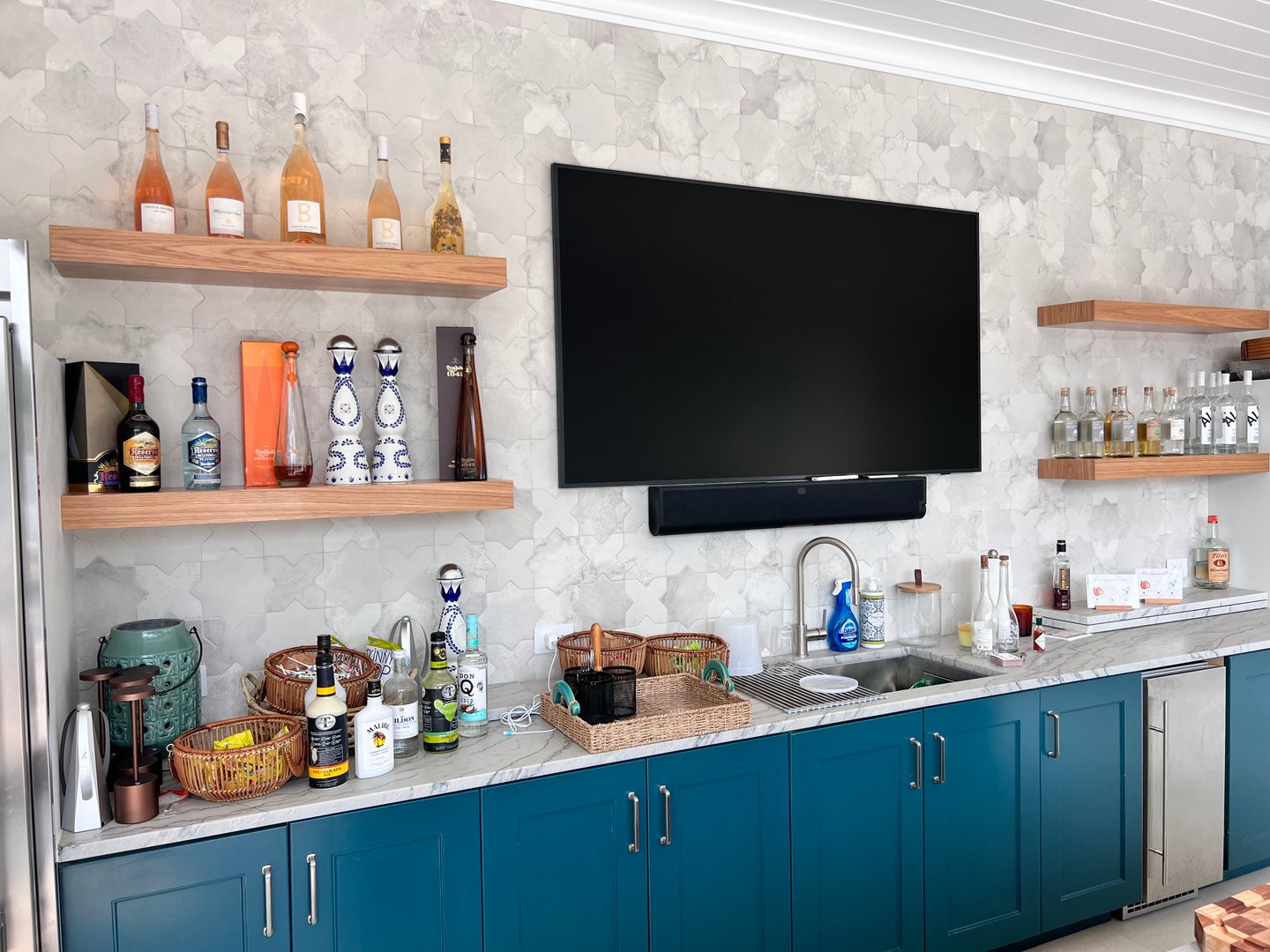Home bar with blue cabinets, a TV, liquor bottles on shelves, and a marble backsplash.