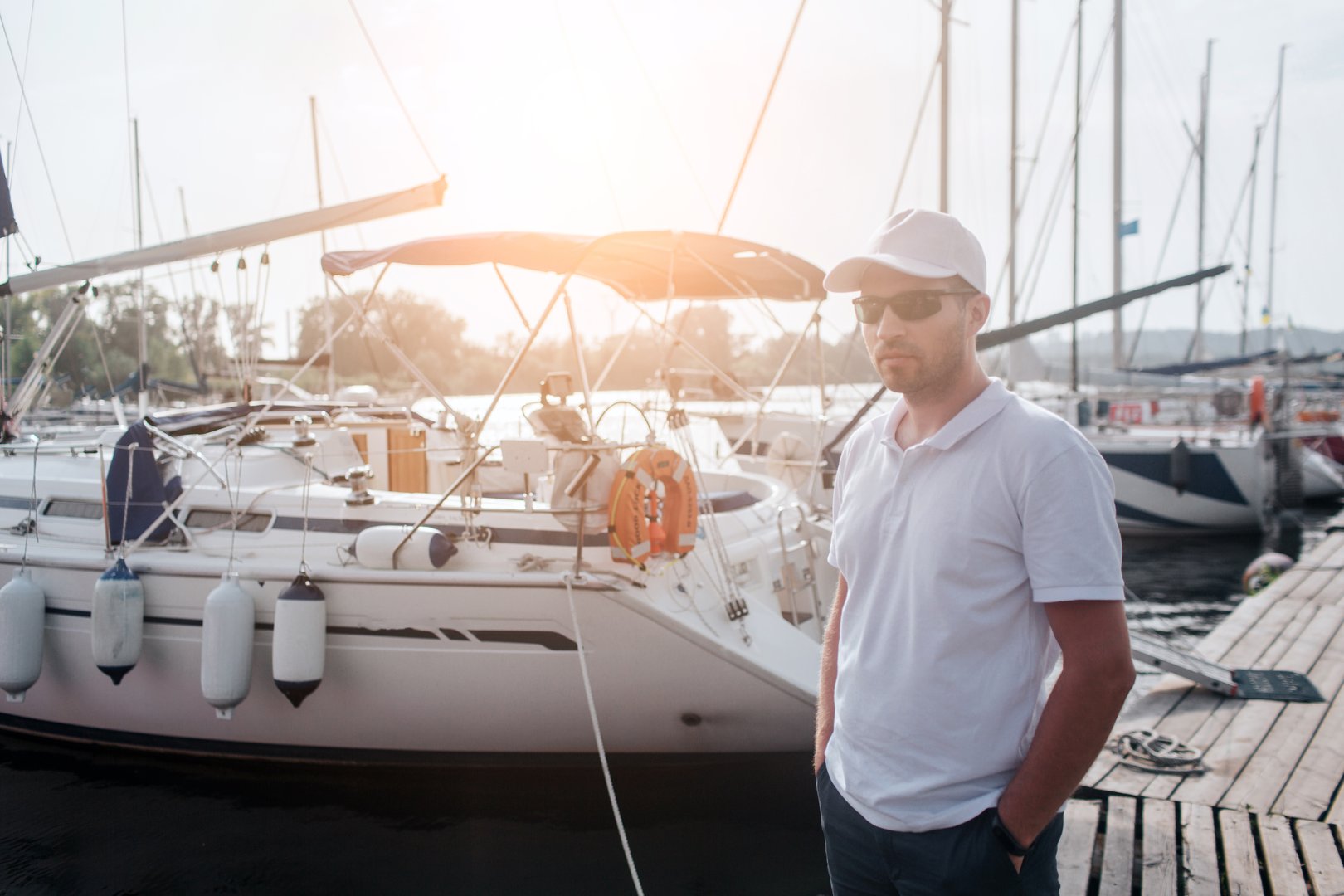 Serious man stands and poses. He holds hands in pockets. Guy look forward, He wears sunglasses, white cal and shirt