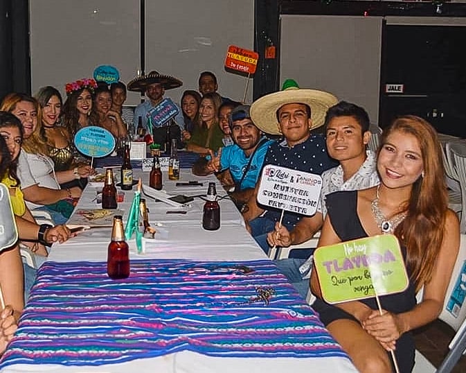 Group of people in festive attire seated at a table with drinks, holding colorful signs, and wearing sombreros.