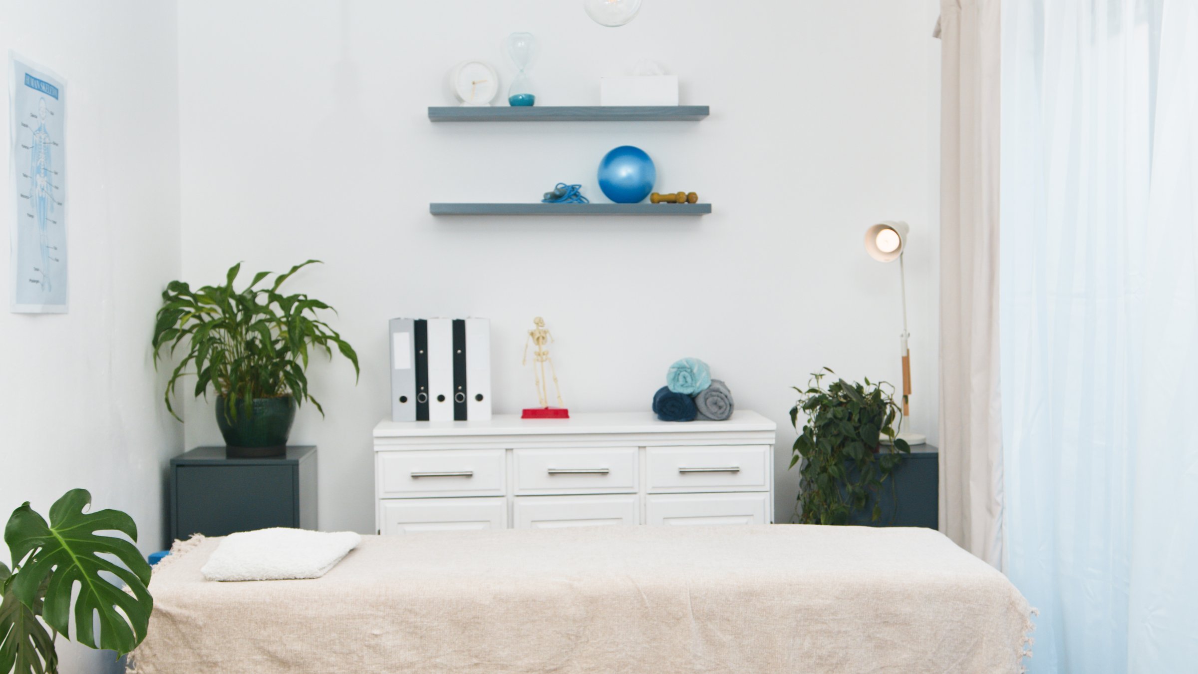 A bright and organized physiotherapy clinic room for patient consultations and rehabilitation. The clean environment features a comfortable treatment bed, shelves with exercise equipment, and anatomical models, well-equipped small practice