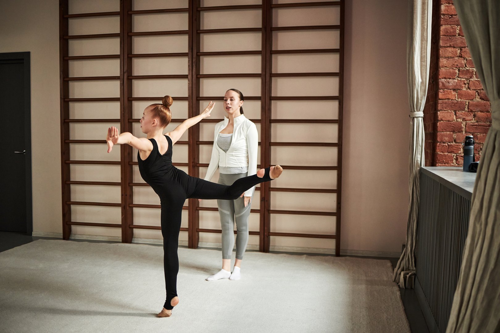 Caucasian girl practicing ballet pose in dance studio while young female coach or instructor observing and giving guidance, wooden wall bars and large window in background