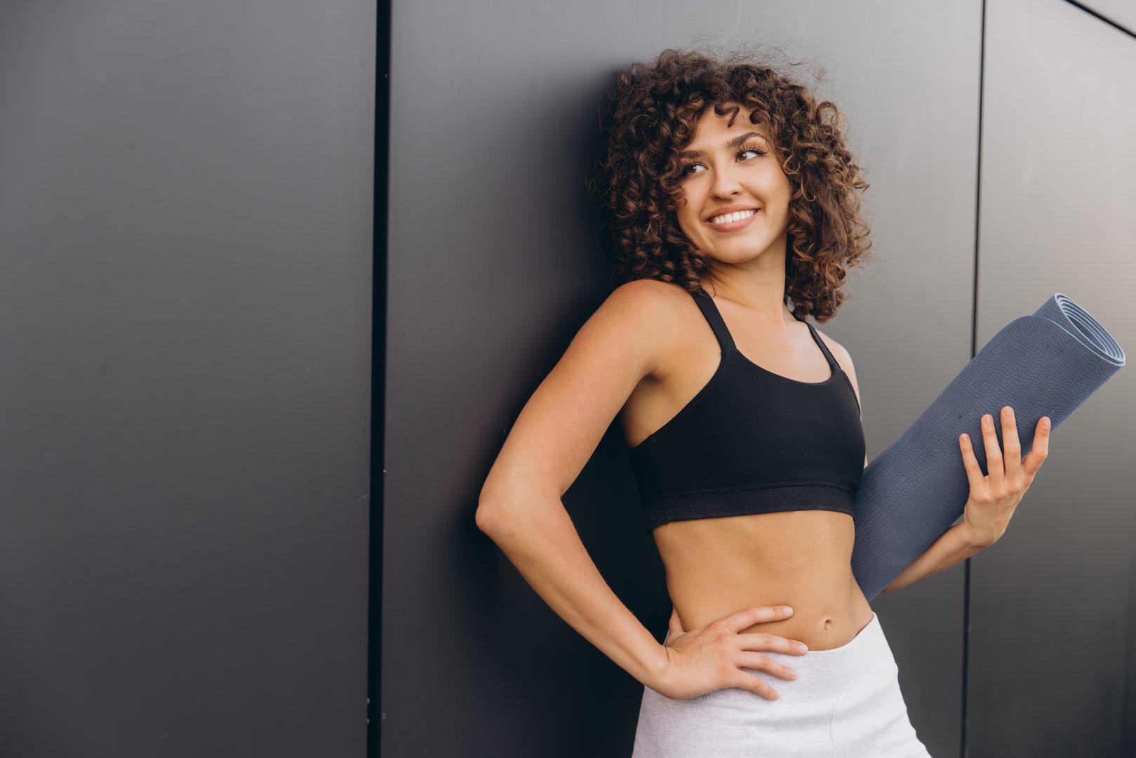 Smiling sportswoman with curly hair, holding a yoga mat while leaning against a black wall, embodies a vibrant and active lifestyle