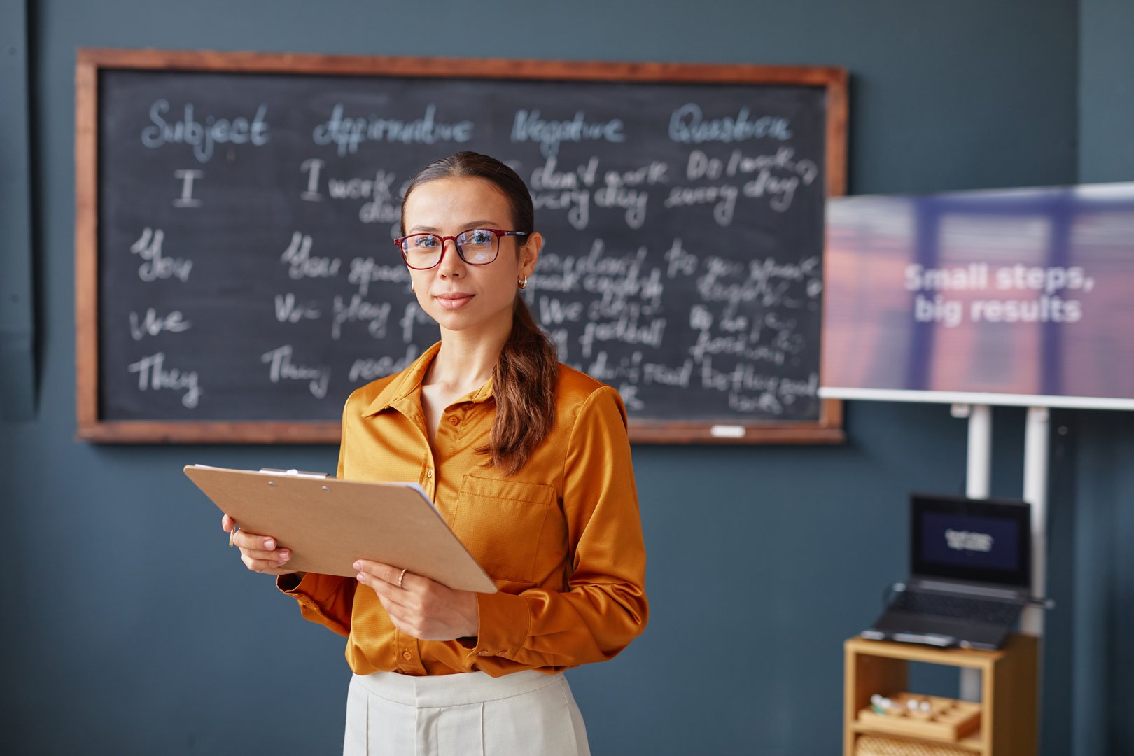 Portrait of young adult Caucasian woman wearing glasses standing in language school classroom holding clipboard, blackboard with English grammar notes and digital screen in background