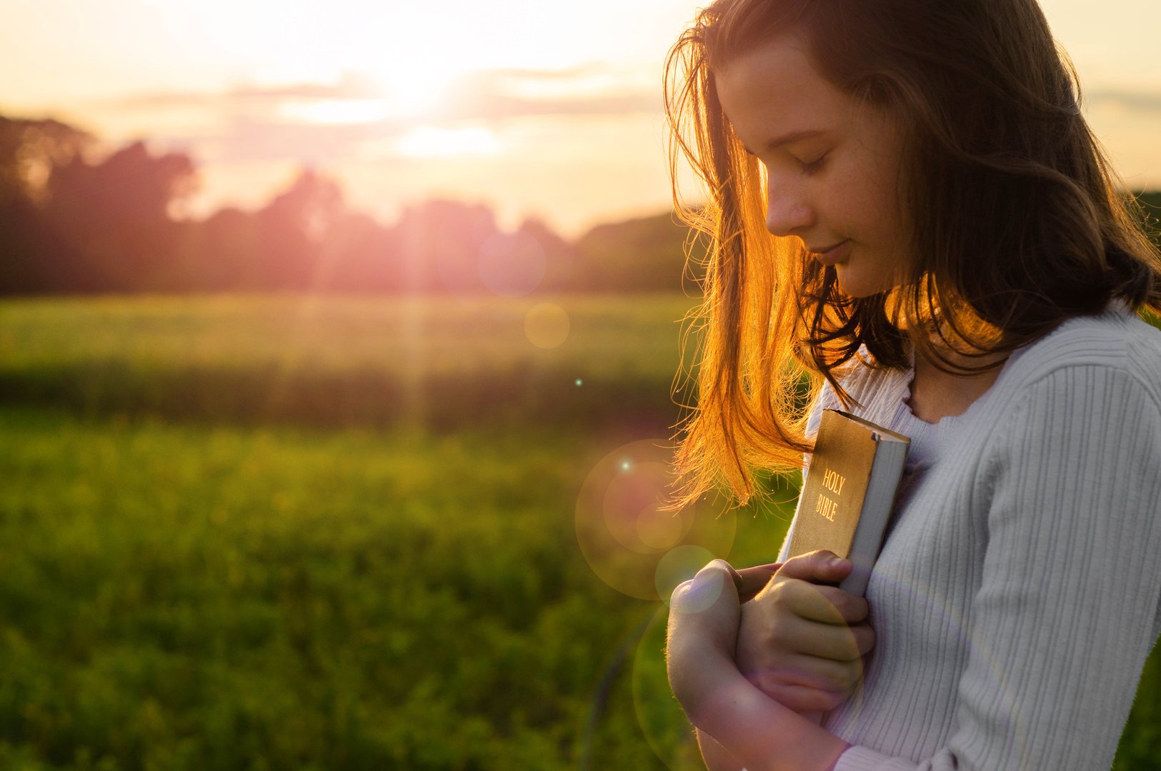 Christian teenage girl holds bible in her hands. Reading the Holy Bible in a field during beautiful sunset. Concept for faith, spirituality and religion. Peace, hope