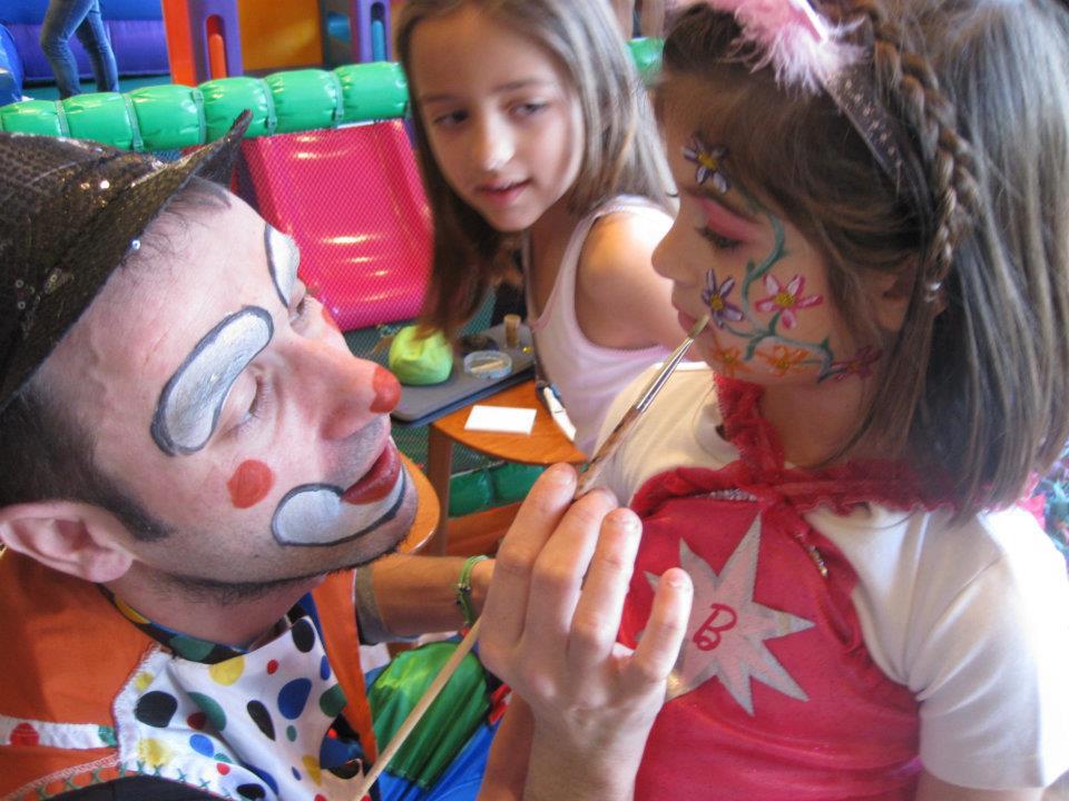 A clown painting a girls face with flowers at a party while another child watches. The girl wears a star-themed outfit.