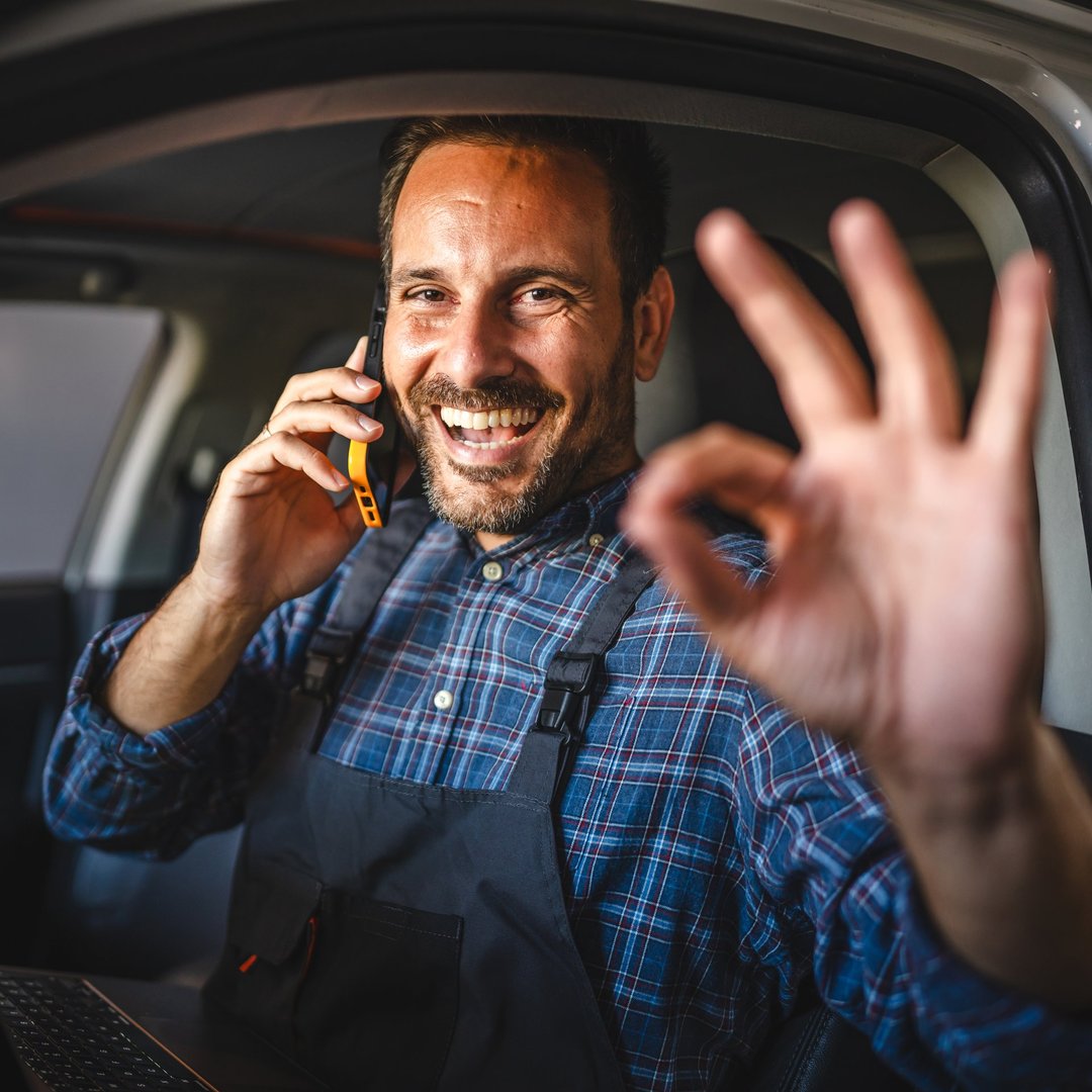 A cheerful car mechanic sit inside a car, hold a mobile phone to his ear with one hand and show an OK sign with the other, expressing satisfaction and success