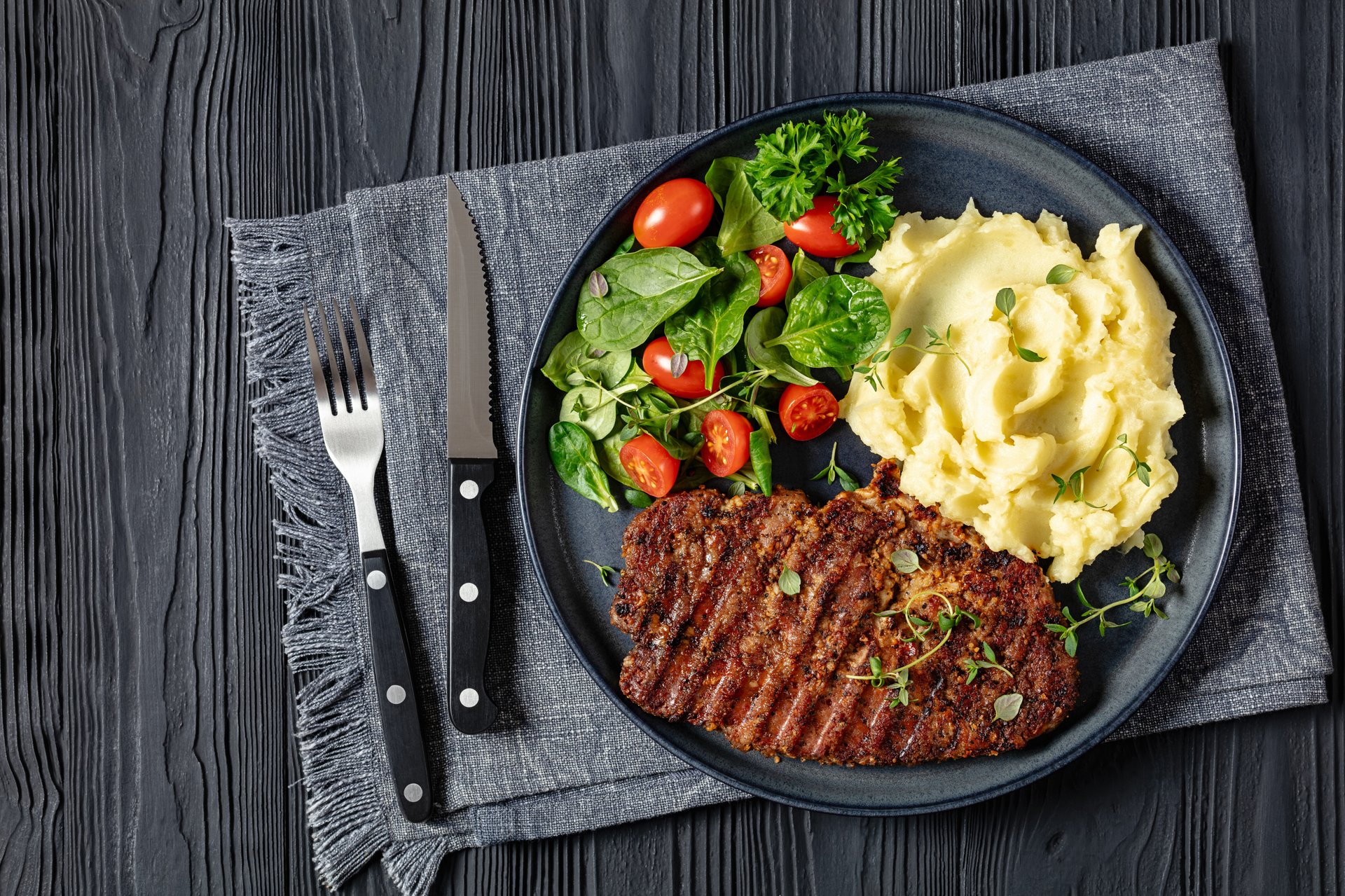 juicy grilled pork steak marinated in spicy sauce served with mashed potato and spinach tomato salad on plate on black wooden table with cutlery, flat lay, free space