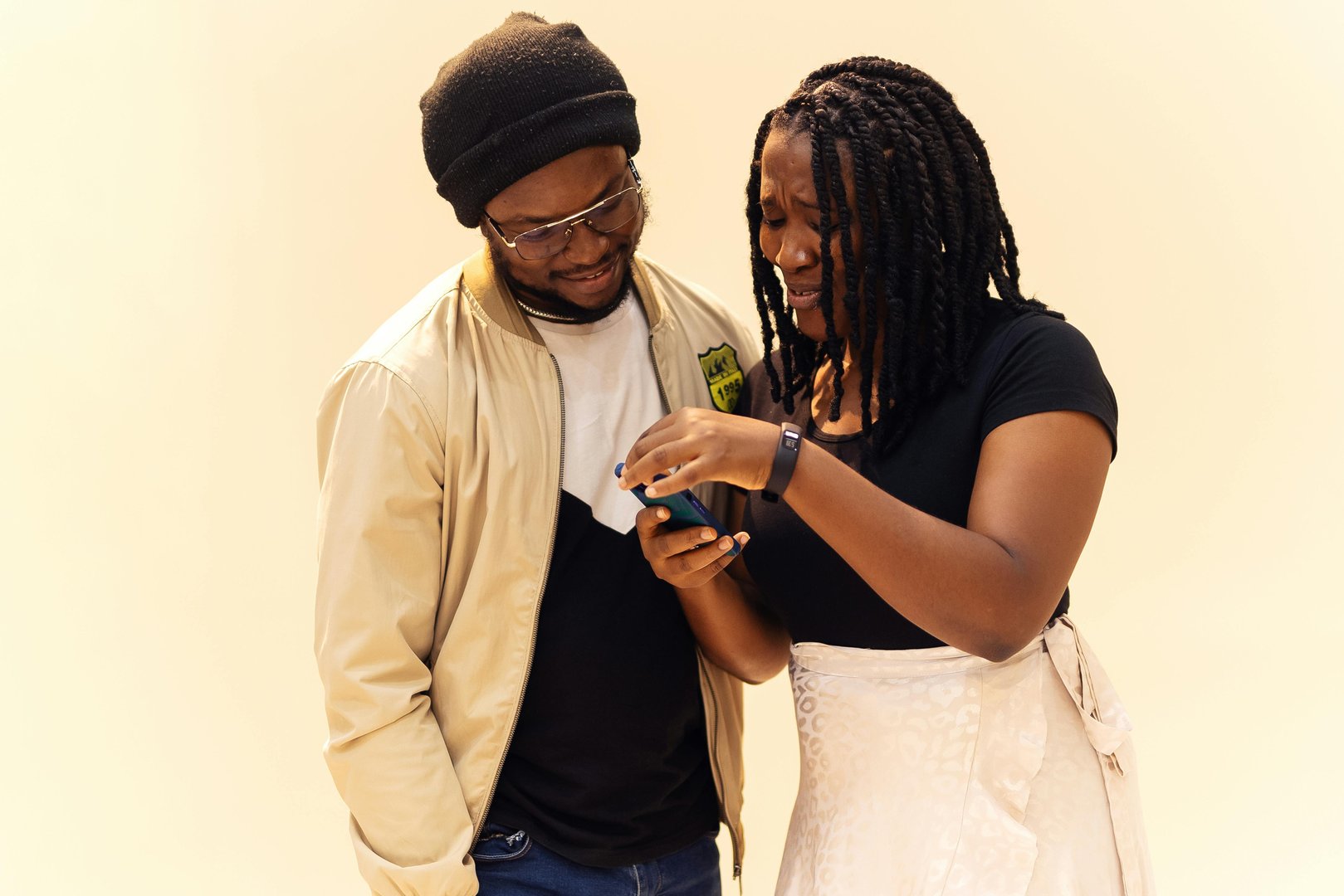 A man and woman smiling while looking at a smartphone together. The woman points at the screen. Beige background.