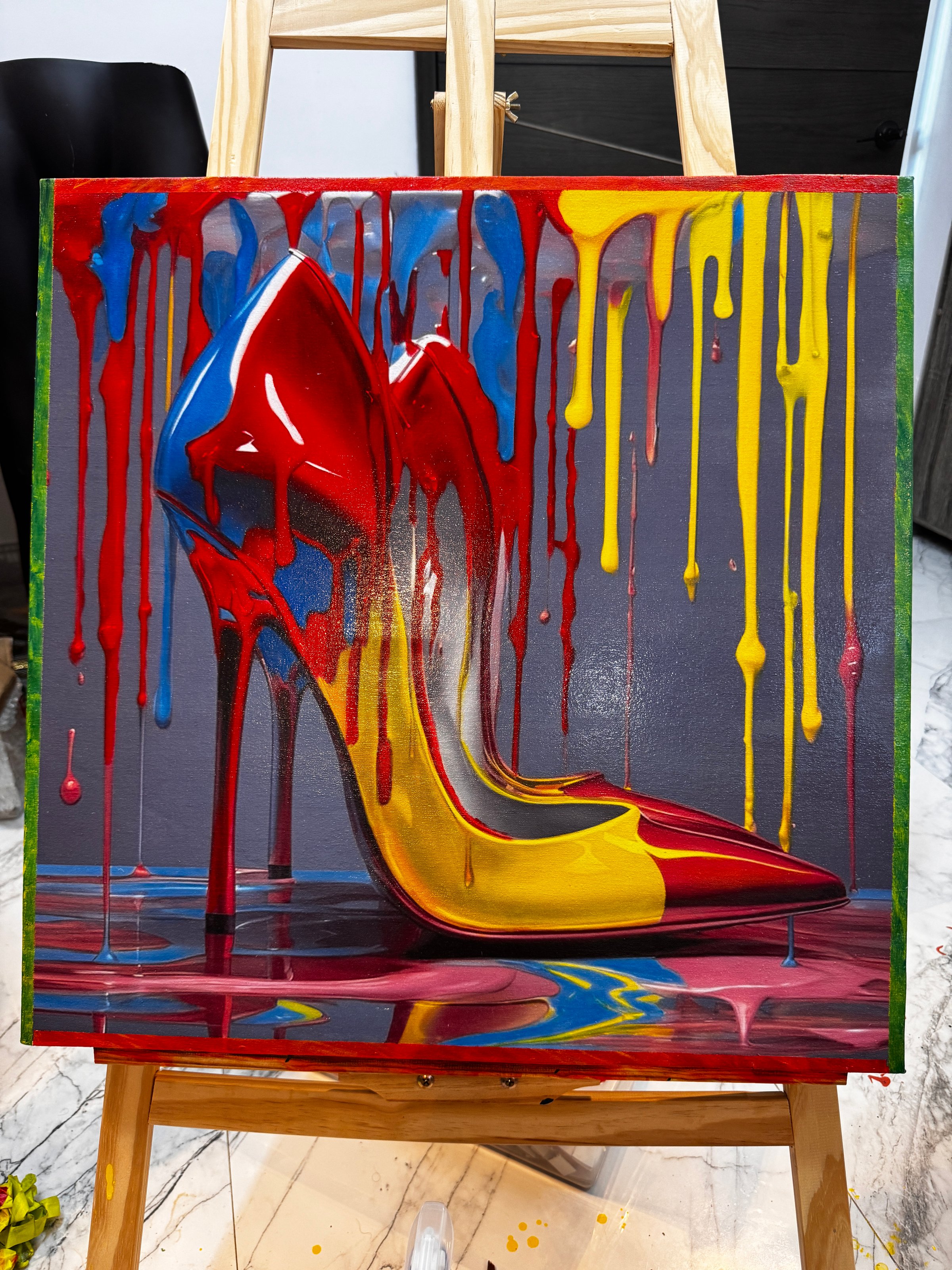 Colorful painting of high-heeled shoe with red, yellow, and blue paint drips on a canvas on an easel.