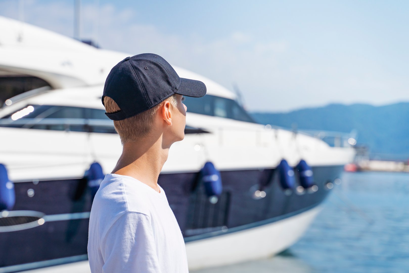 Teenager dreams, looks to the future on the marina. Teen boy in seaport with sailing ships young man is wearing a black baseball cap and white t-shirt  Sailing vessels drop anchor in a quiet bay