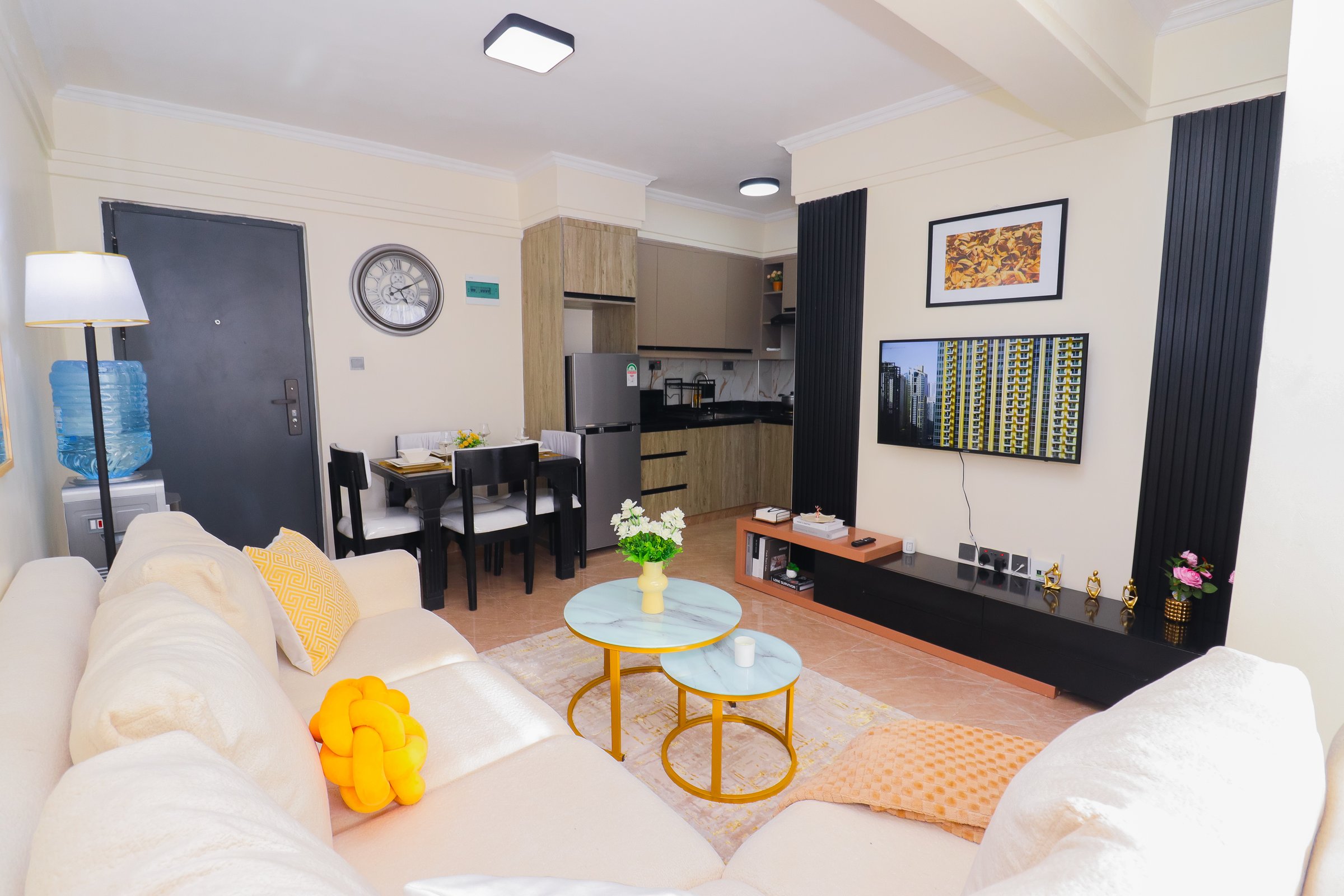 Modern living room with beige sofa, round tables, TV, and dining area beside compact kitchen. Decor includes plants and artwork.