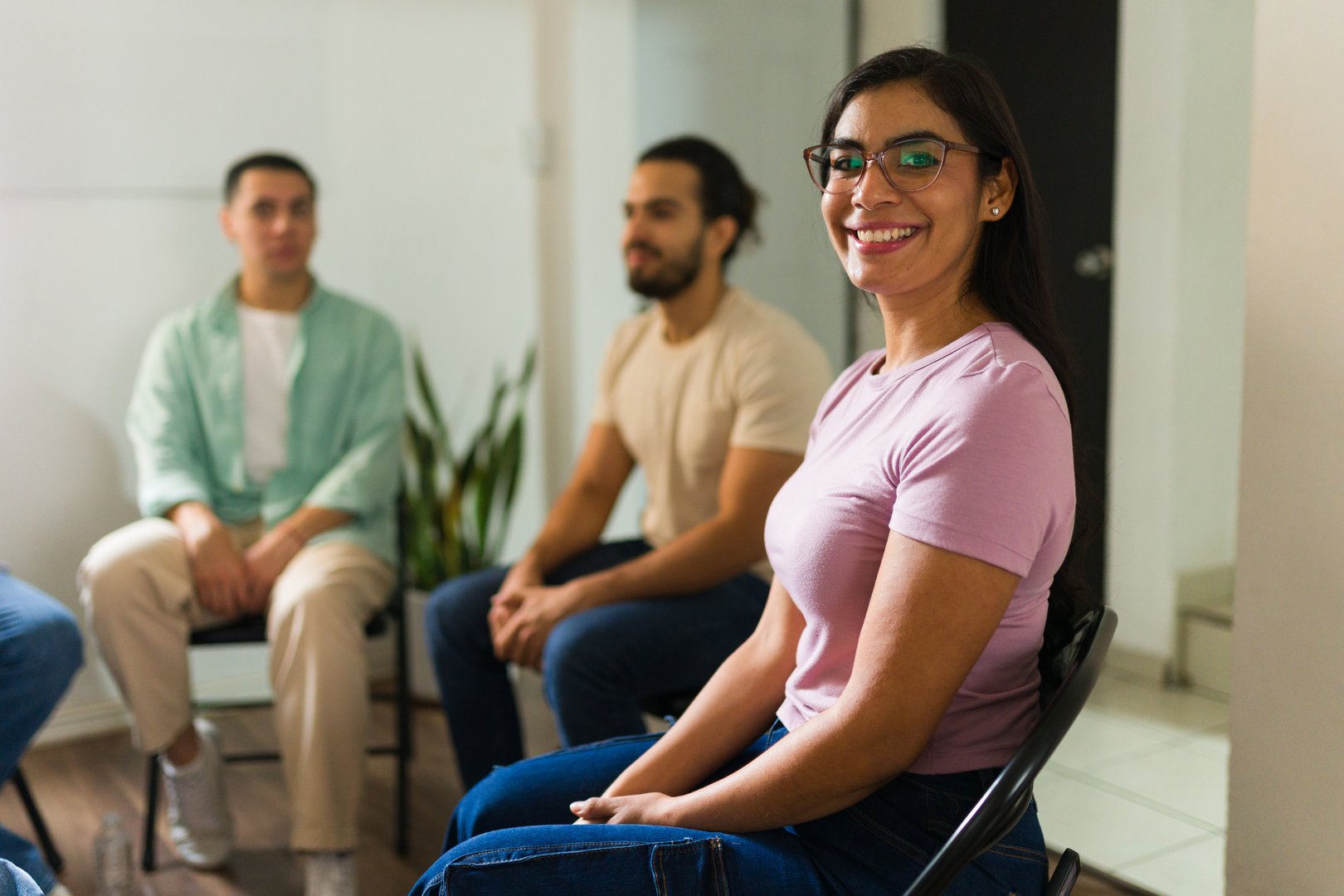 Woman in glasses smiling at camera during a support group meeting. Diverse people sitting on chairs, receiving mental health therapy
