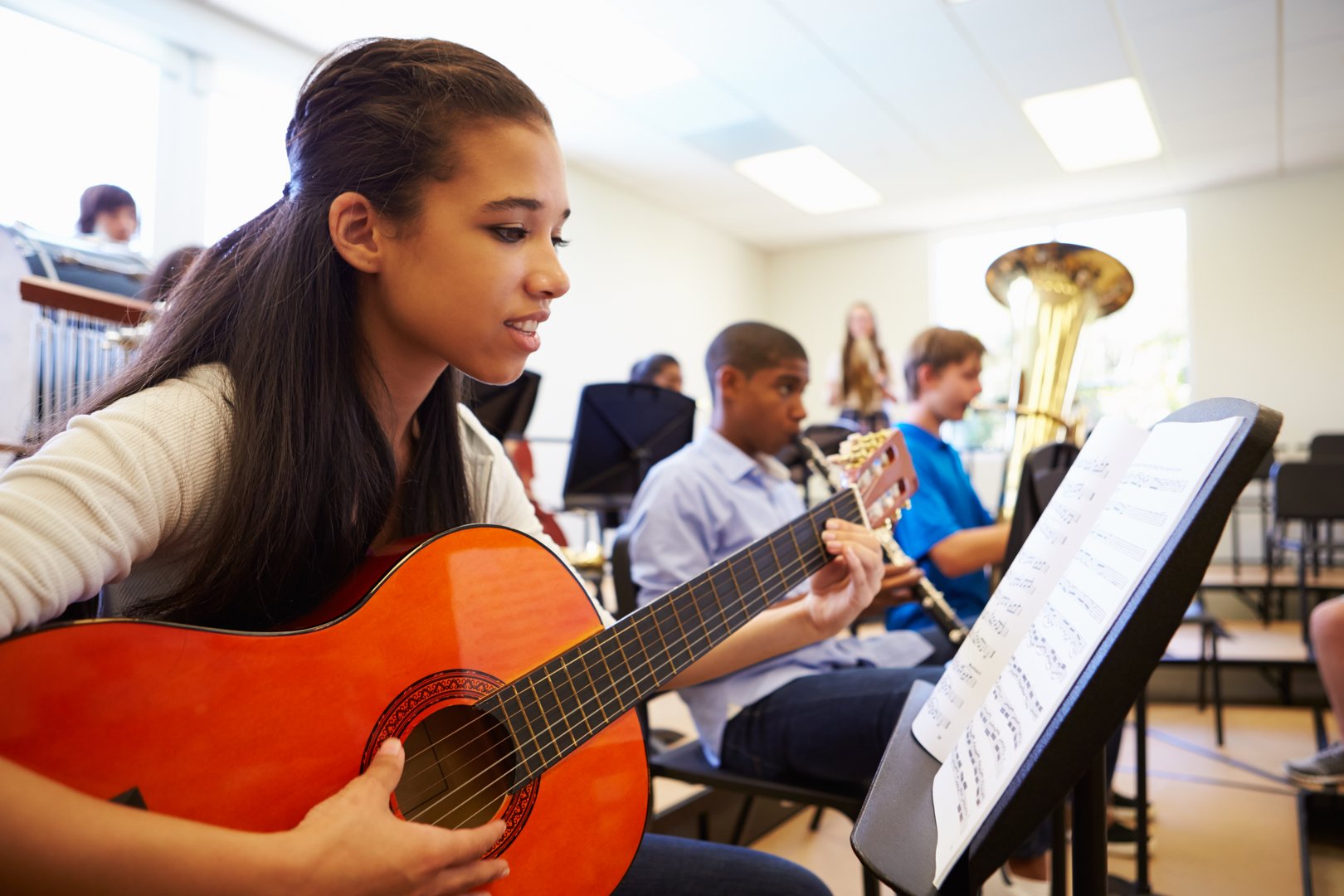 Female Pupil Playing Guitar In High School Orchestra Reading Music Sheet