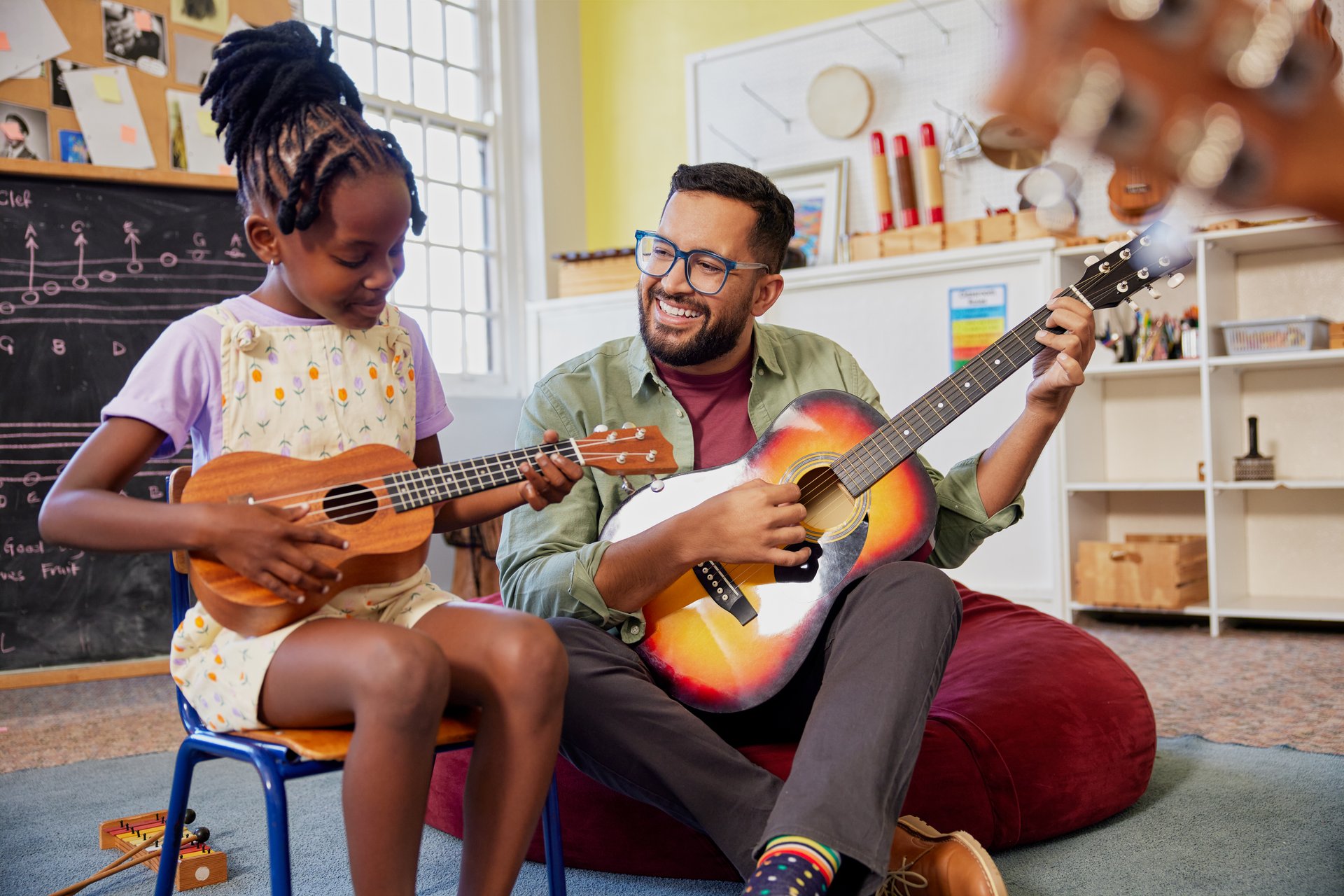 Happy male teacher assisting an african kid to play musical instrument in classroom at school. Happy teacher playing acoustic guitar and singing while having music class with girl. Cute little girl learn to play ukulele with male teacher.
