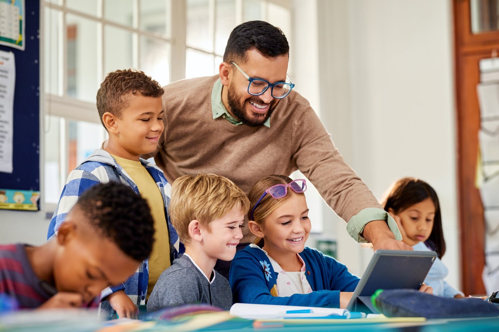 Cheerful male teacher helping students using digital tablet in classroom during technology lesson. Helpful male professor talking to multiethnic students using digital tablet in classroom. Male teacher supervising a group of elementary children working on computer.