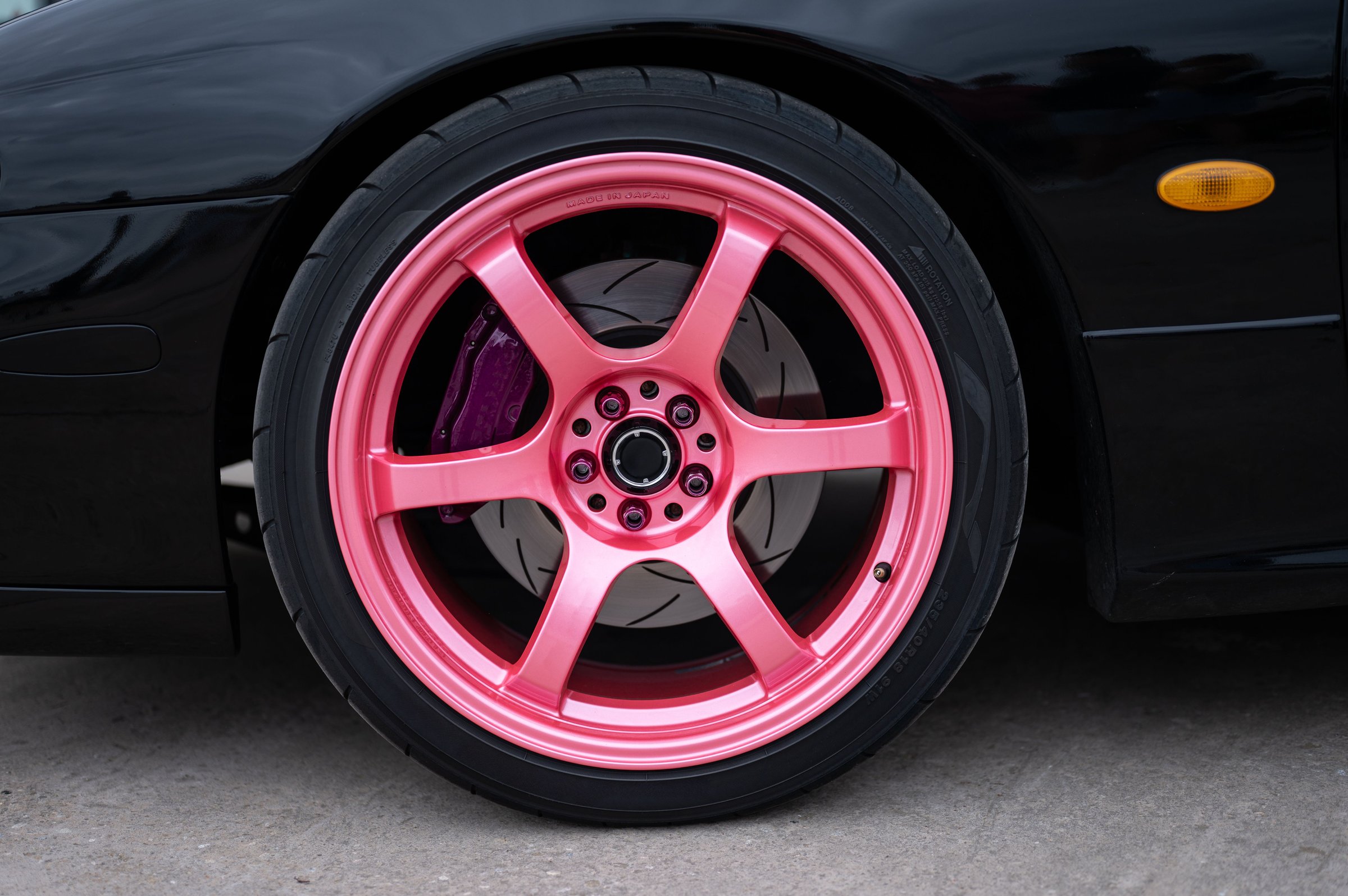 A detail of the front wheel on a JDM street racing and drift sports car, featuring a pink Gram Lights wheel and a modified black brake system.