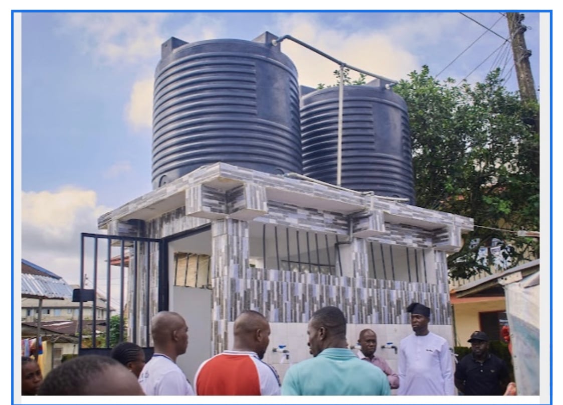 People gathered near a building with two large water tanks on the roof. The structure has tiled walls and is outdoors.