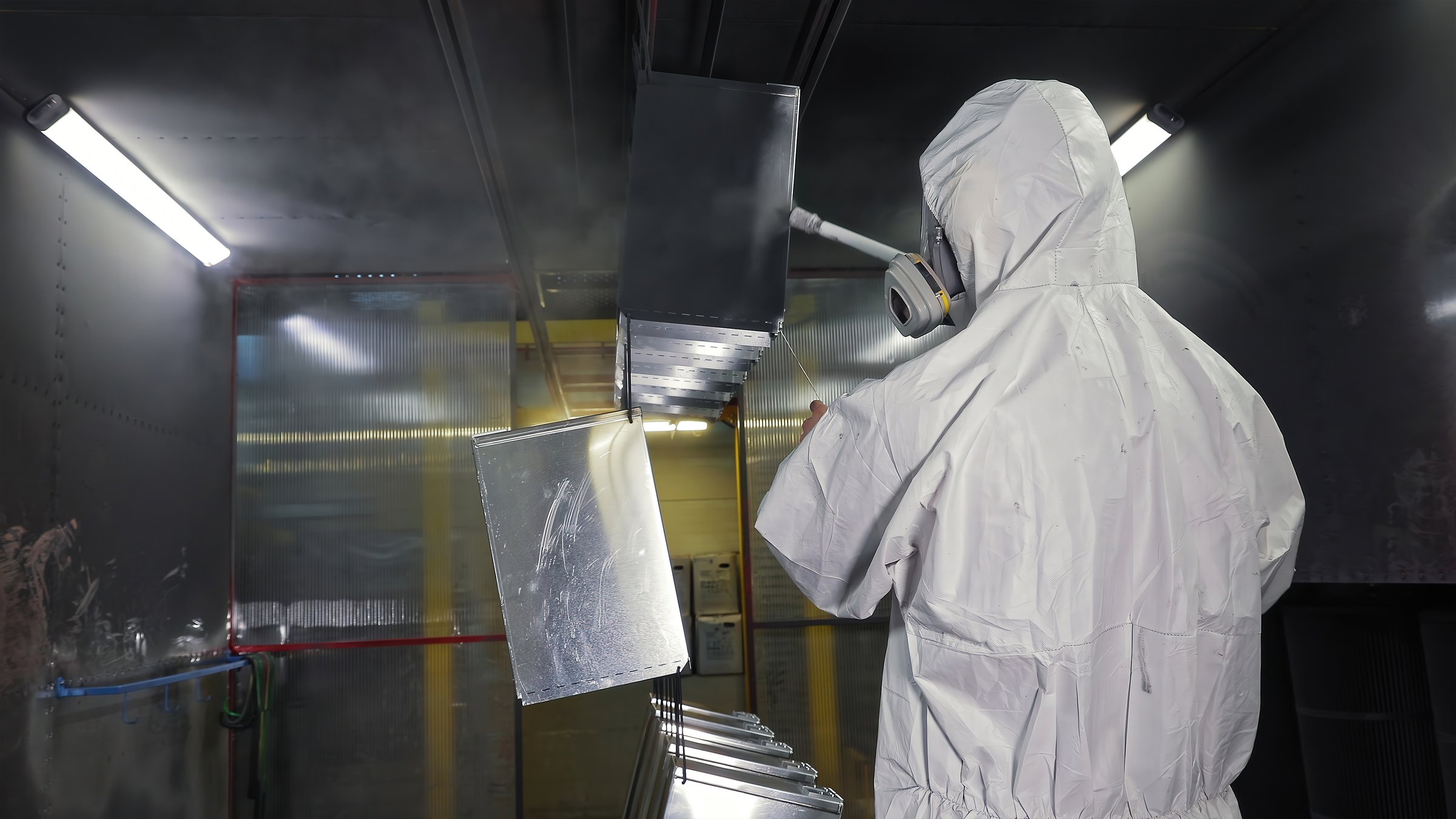 Worker in protective suit spray-painting metal parts. Industrial worker in hazmat suit using a spray gun to paint metal components inside a controlled spray booth environment