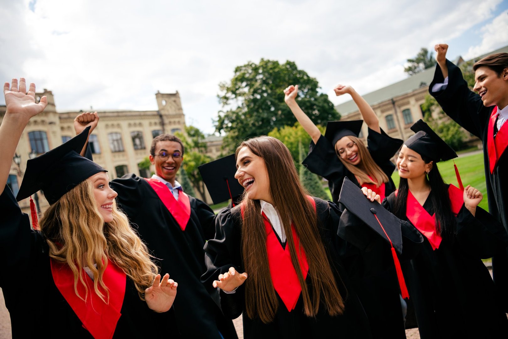 A group of happy young graduates celebrate outside a university, wearing caps and gowns. They cheer together on a sunny day, marking their academic success.