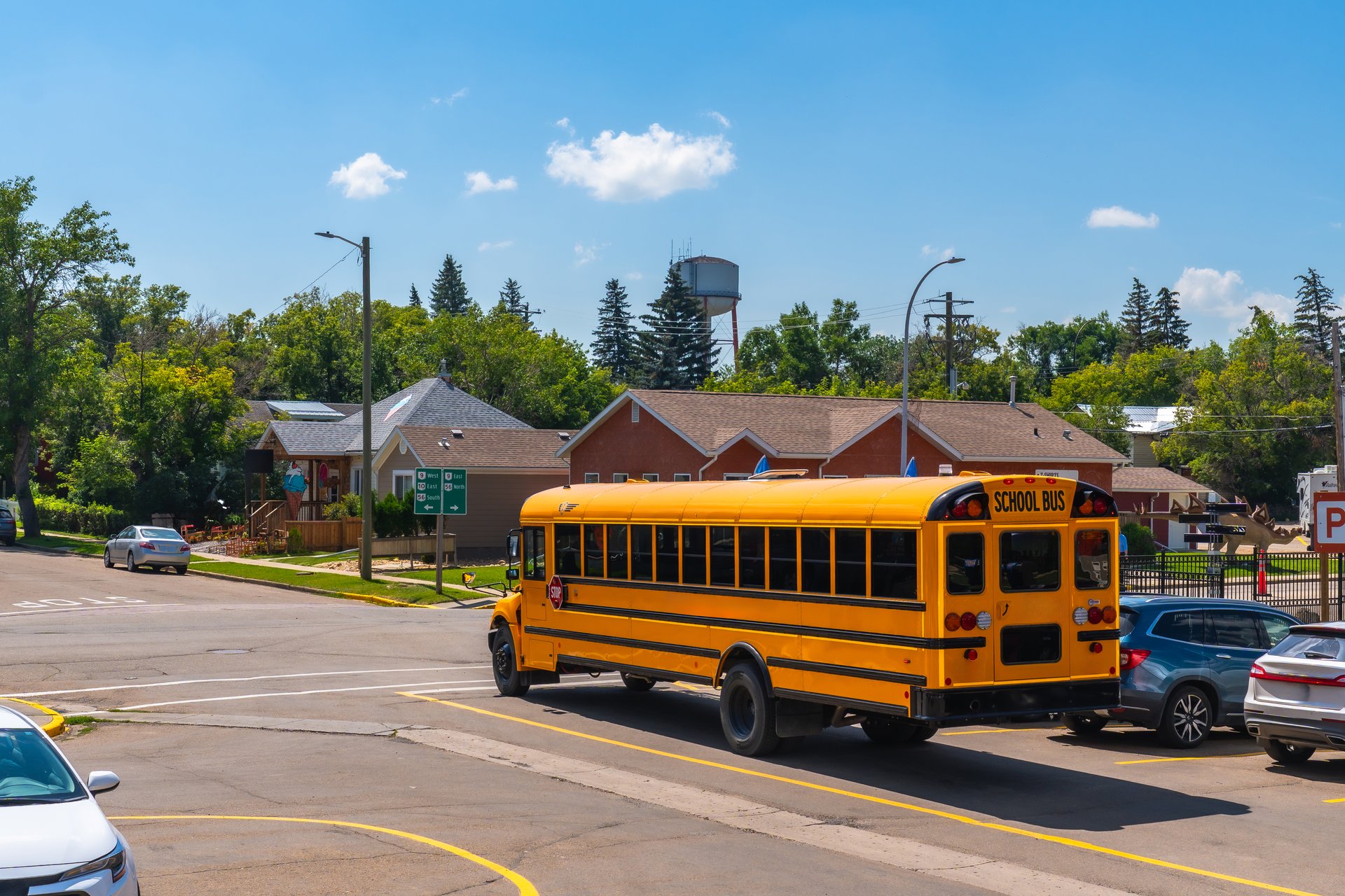 Yellow school bus driving on a street in a residential neighborhood, leaving a parking lot and passing by houses and trees on a sunny summer day