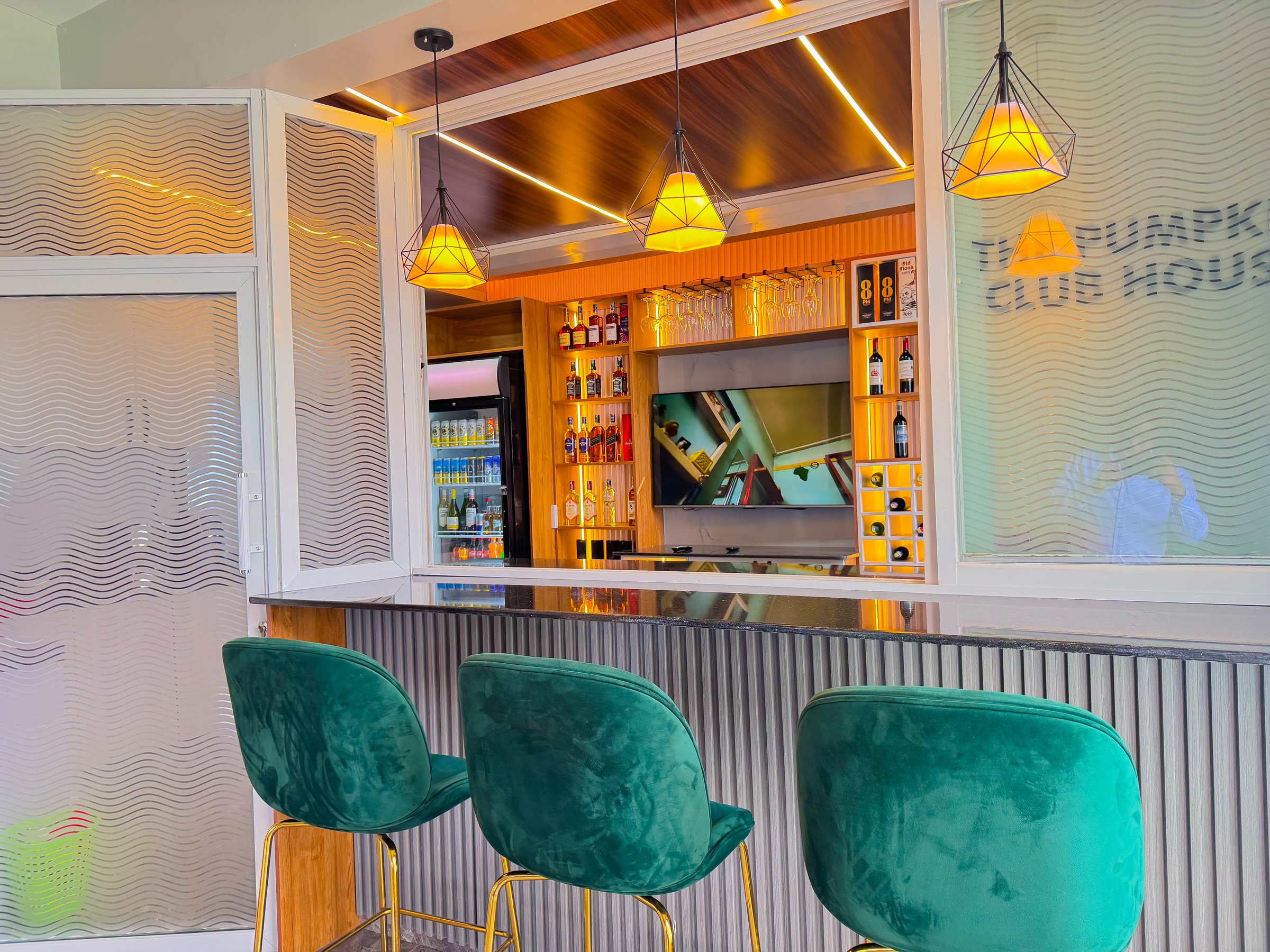 Chic bar area with green velvet stools, a wooden counter, and backlit shelves displaying various bottles and a TV screen.