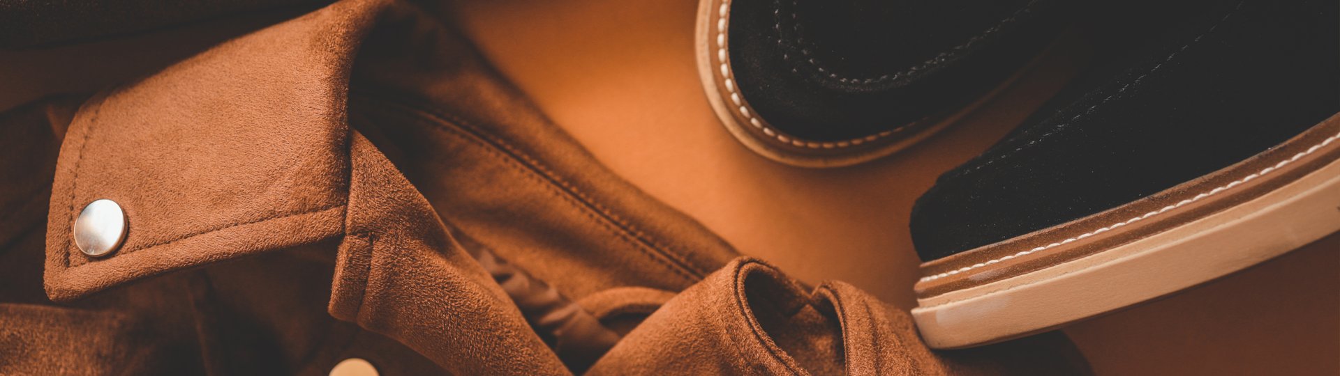A wide, minimal, and modern header featuring a brown suede jacket and black shoes, perfect for a website background with copy space.
