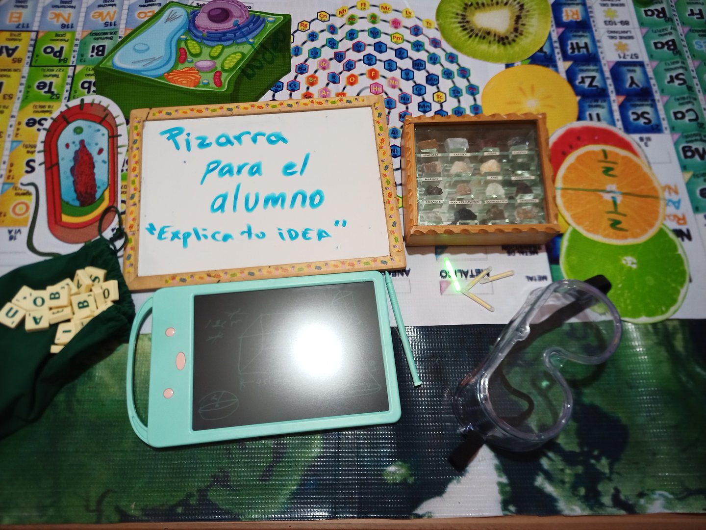 Educational table with a mini chalkboard, small tablet, chemistry kit, letters, and goggles for a classroom activity.