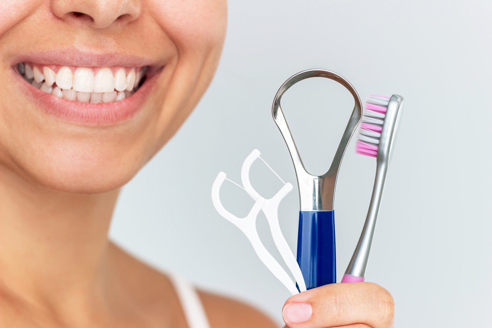 Close-up of a young smiling woman holding dental care tools: toothbrush, tongue scraper, and dental floss picks on a light background. Concept of morning hygiene, oral care routine, and healthy habits