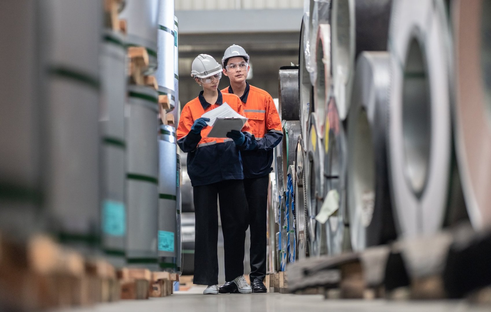 A male and female engineer perform a meticulous inventory audit, walking through the storage area of a metal sheet factory, ensuring stock accuracy and production readiness