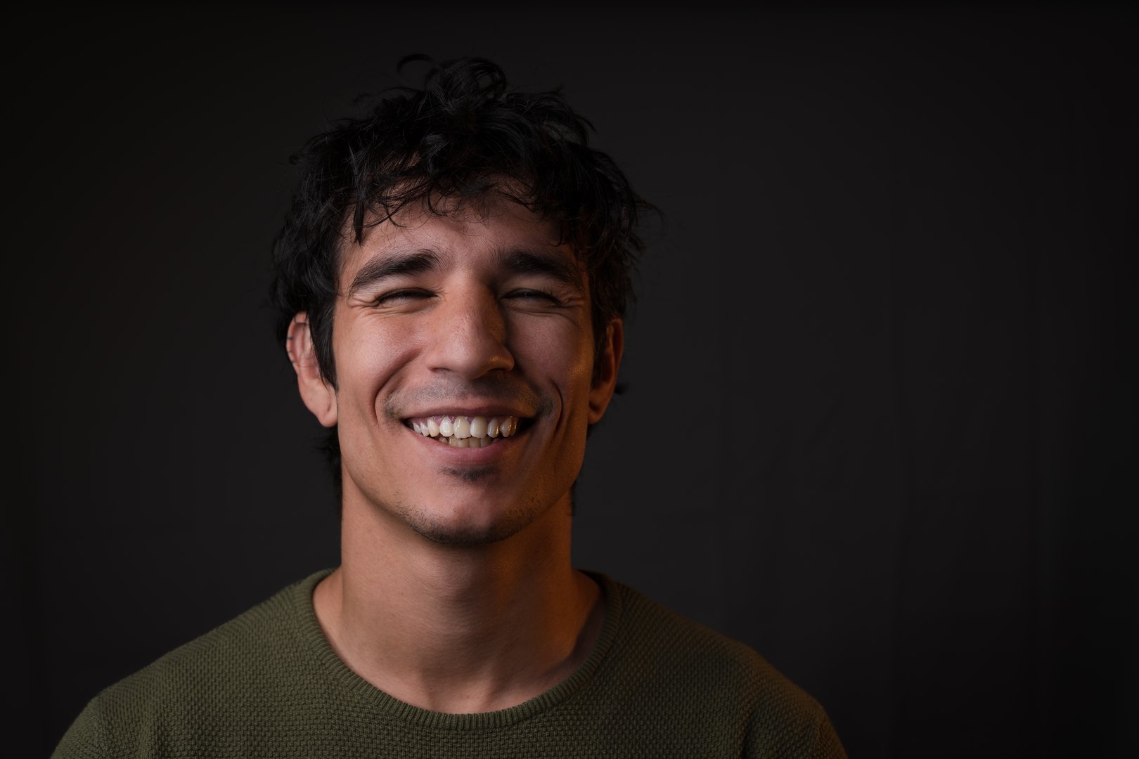 A radiant portrait of a young Latino man caught in a burst of genuine laughter, his face beautifully illuminated by contrasting lighting. A crisp white sidelight highlights his joyful expression while a golden opposing light creates dimension, casting a warm glow on his skin and catching playful highlights in his hair. The deep black background makes his vibrant energy pop, resulting in a perfect balance between professional polish and infectious happiness. This dynamic image is ideal for representing positivity, successful communication, or cultural diversity in business and lifestyle contexts.