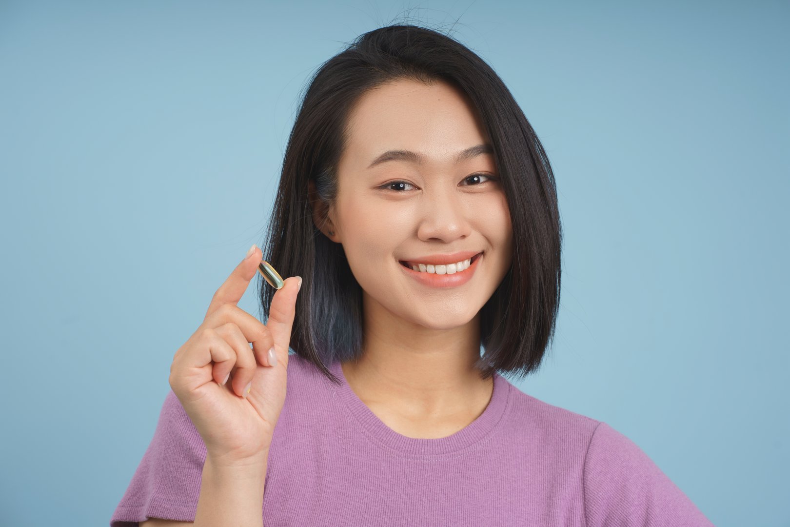 A young Asian woman with short black hair wearing a purple shirt is smiling while holding a small object between her fingers. The bright blue background adds a cheerful vibe to the moment.