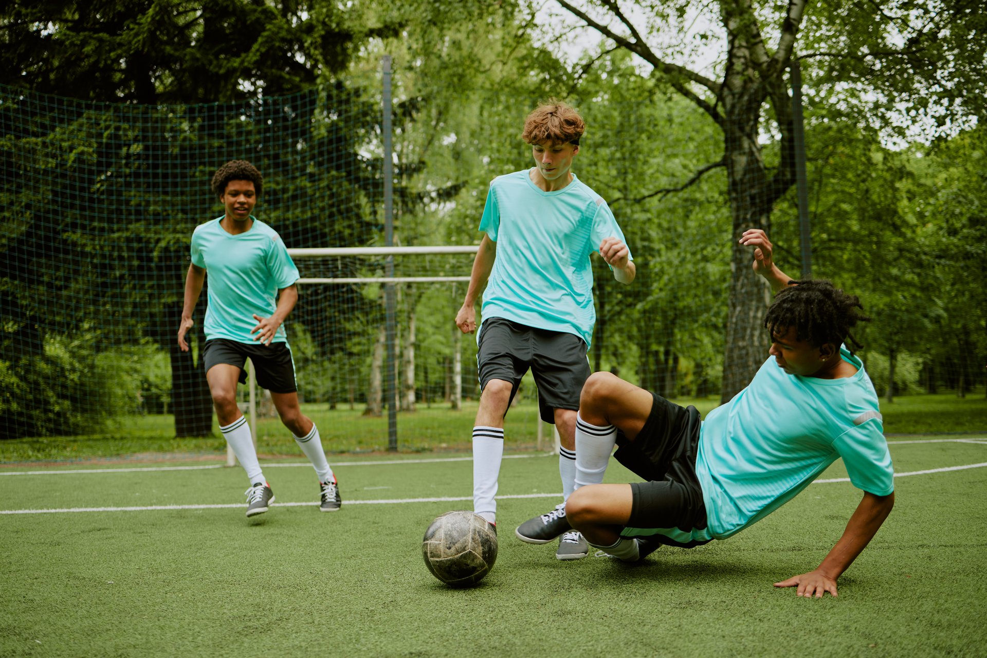 Three teenage boys playing soccer on outdoor field, one Black teenager sliding to tackle ball while others running and focusing on game action