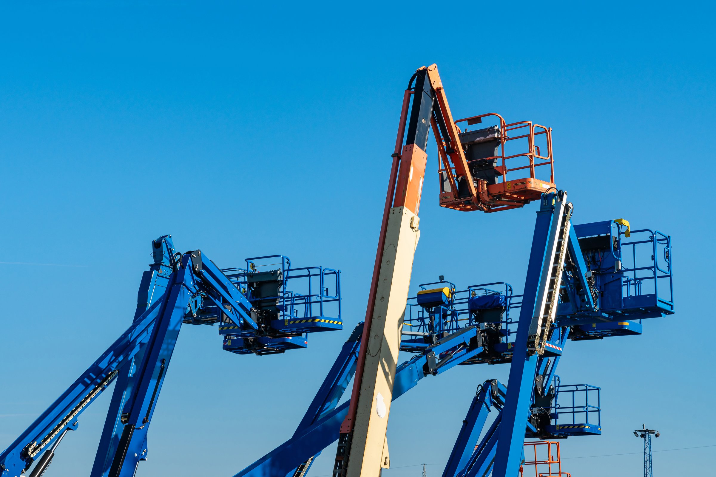 Multiple boom lifts in various colors and heights, reaching towards the sky, set against a clear blue background at a construction site.