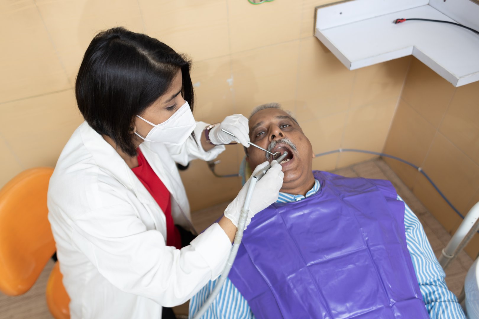 Female dentist in gloves and mask examines an elderly male patient in a modern dental chair.