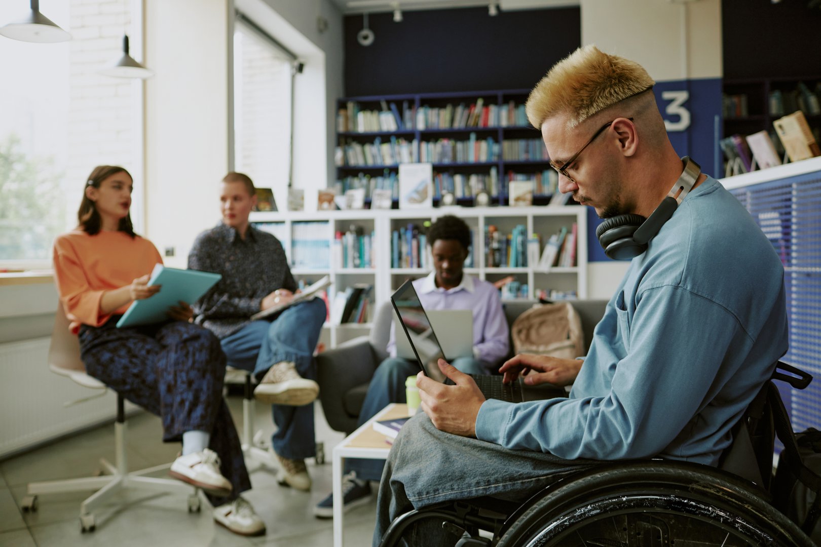Group of diverse individuals engaging in study session in modern library workspace. Man in wheelchair focusing on work with laptop, while others discussing and taking notes