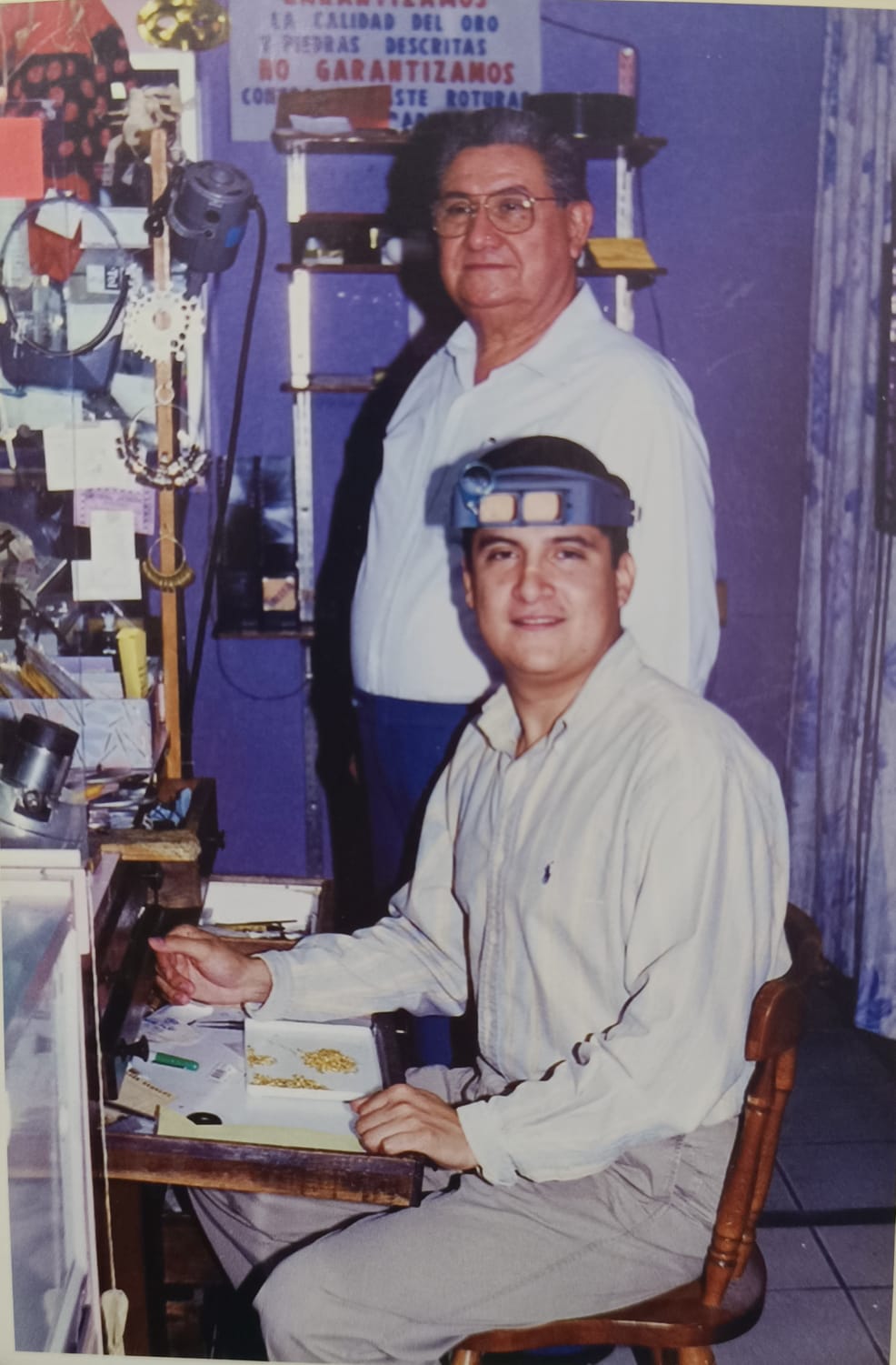 Two men in a workshop; one standing, one seated with magnifying goggles, amidst tools and equipment on shelves.