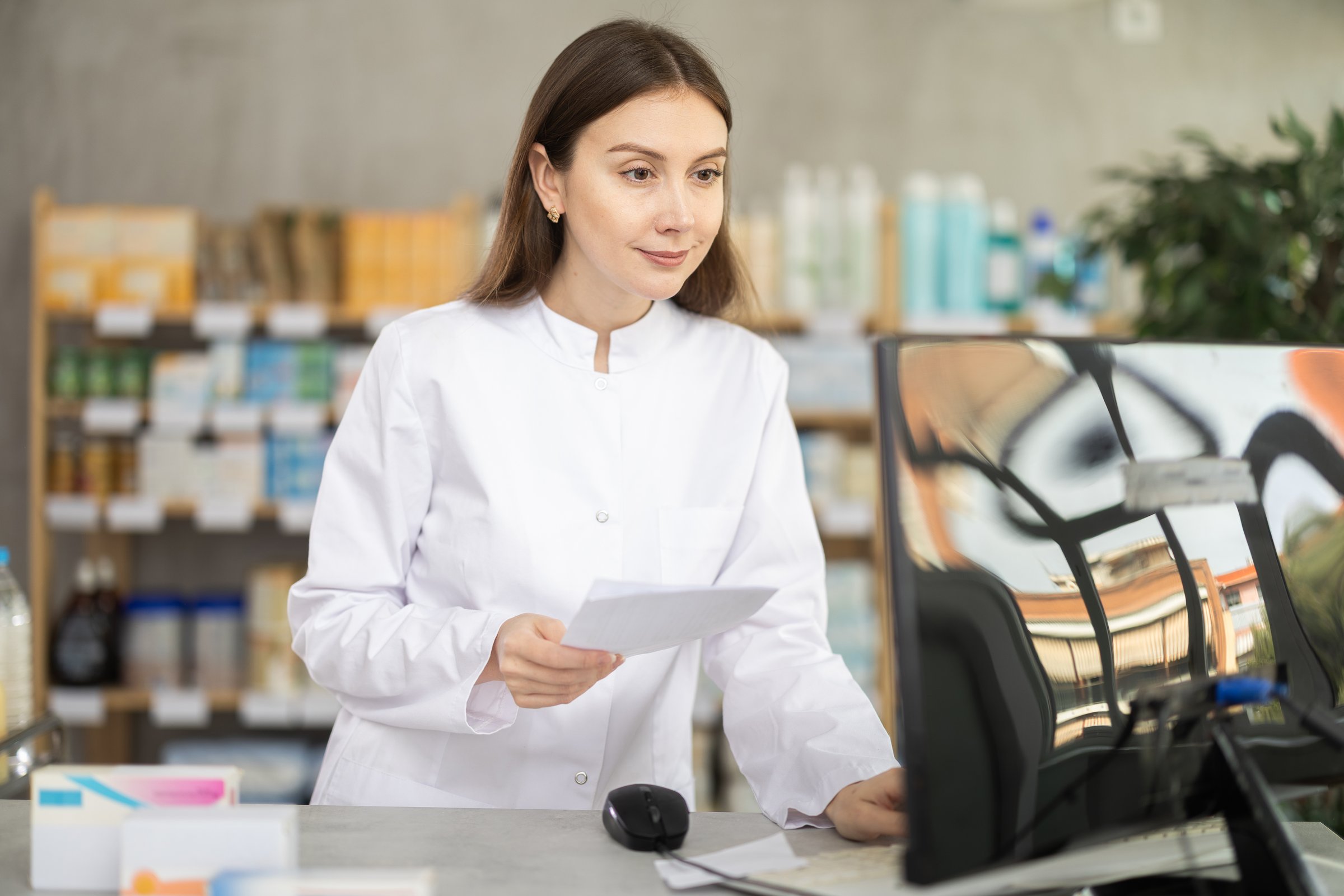 Young woman pharmacist looking for box of pills on computer in pharmacy