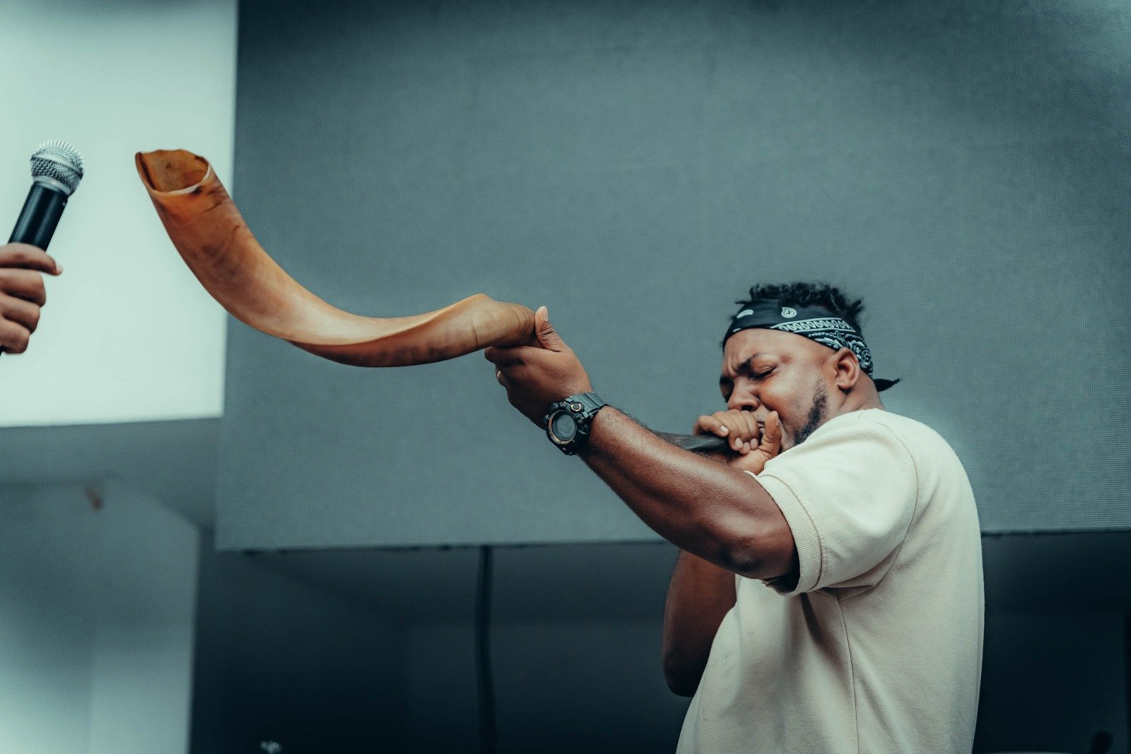 Man blowing a shofar while holding a microphone, with another hand holding a microphone nearby.