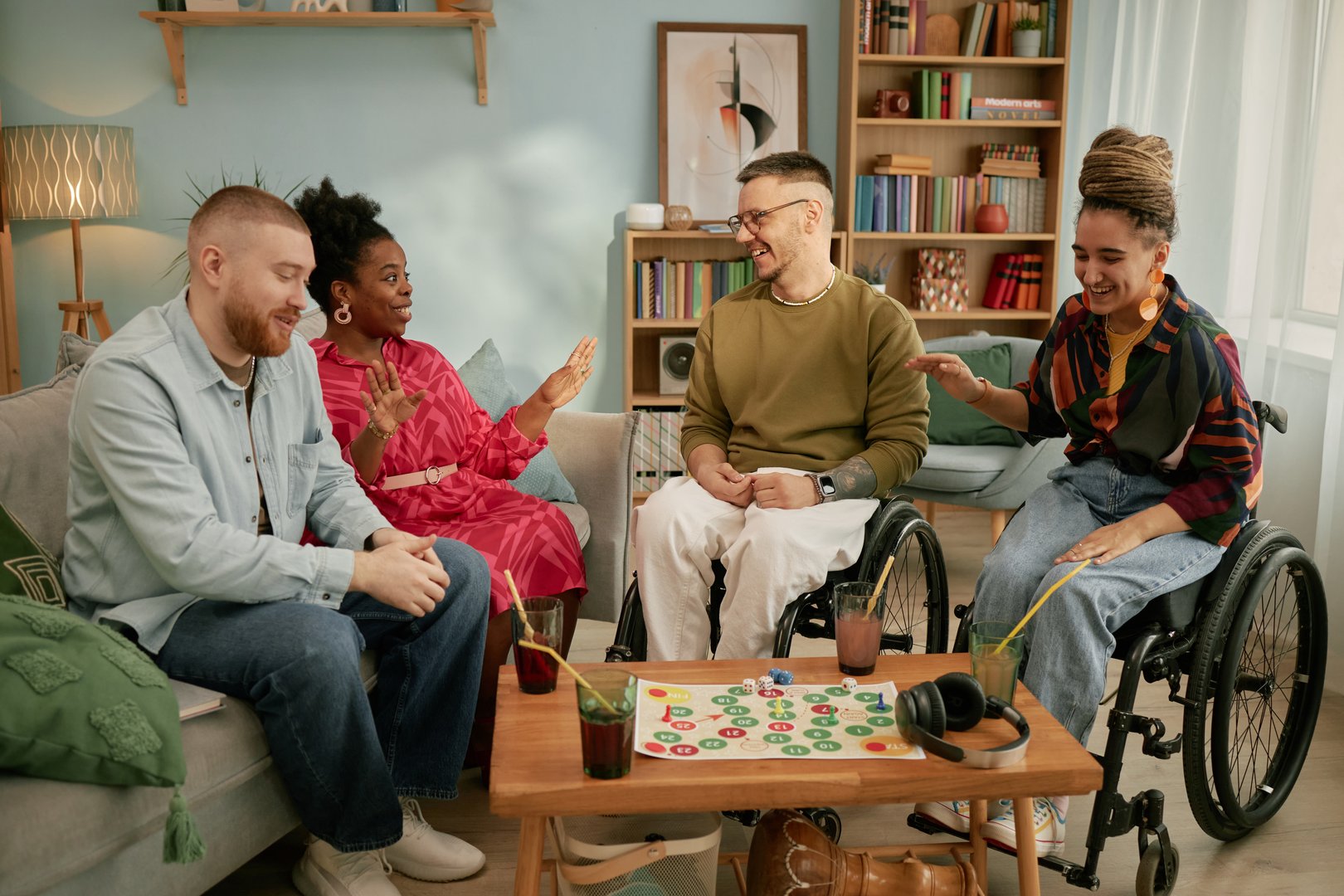 Diverse group of young adults and middle aged men and women with disability sitting together around board game table celebrating birthday, smiling and interacting in living room