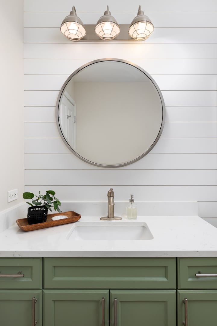 A bathroom detail with a green cabinet, white marble countertop, shiplap accent wall, and a light fixture mounted over a circular mirror.