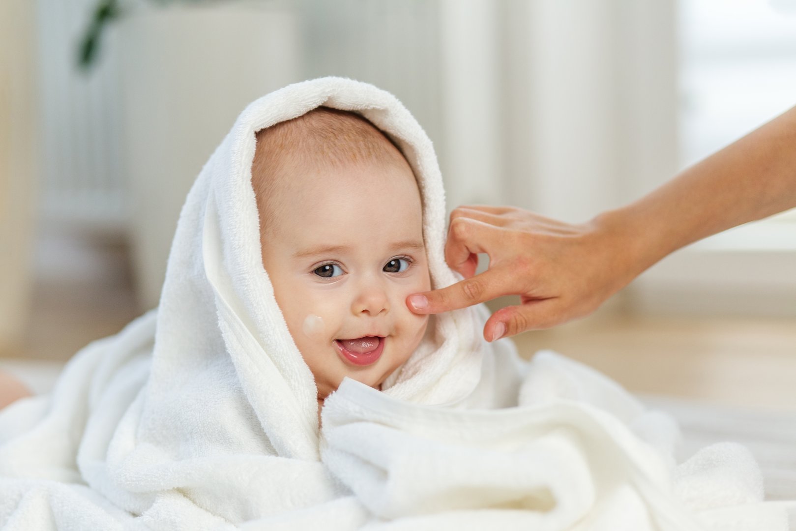 Smiling baby wrapped in white towel receives gentle skincare as cream is applied to the cheek in a cozy home.