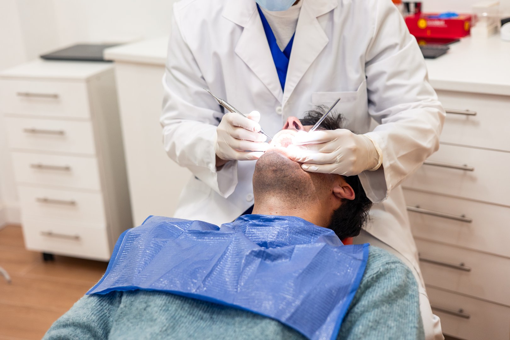 Dentist wearing a mask and gloves is performing a dental check-up on a male patient lying on a dental chair in a brightly lit clinic, using specialized tools and a focused light